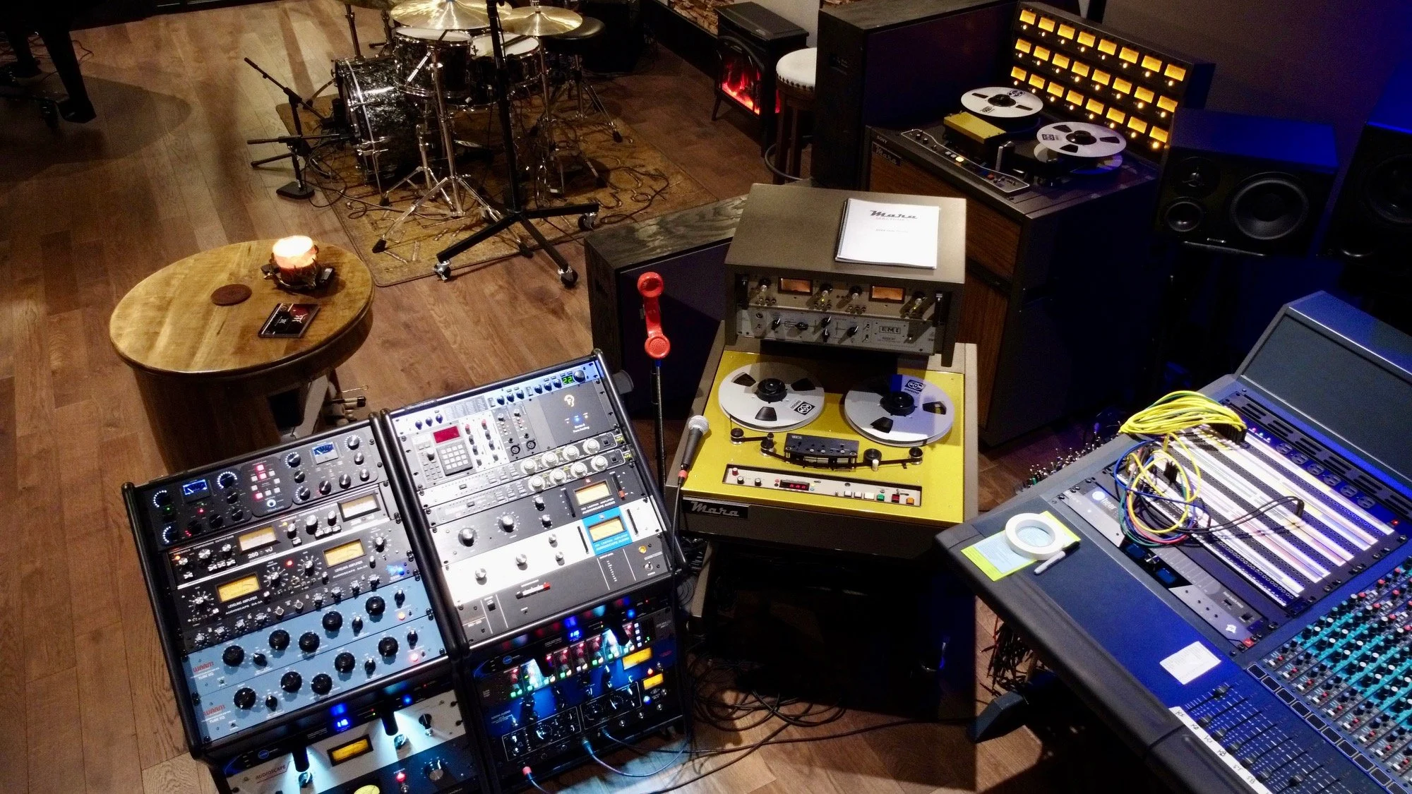 Recording studio with various audio equipment including mixing consoles, reel-to-reel tape recorders, speakers, and drums in the background.