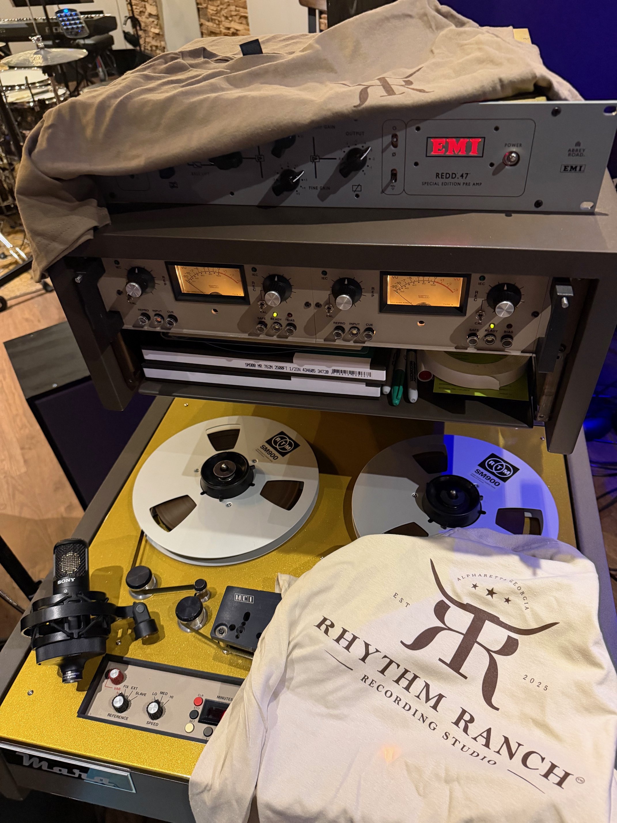 Music recording studio equipment including a pair of reel-to-reel tape decks, a microphone, a microphone stand, and covers or bags with the Rhythmm Ranch logo on them.