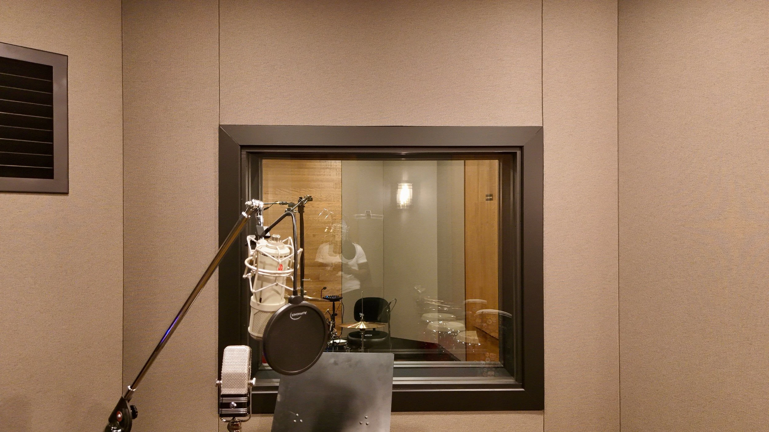 Recording studio with a microphone, music stand, and a stool visible through a window in soundproof wall.