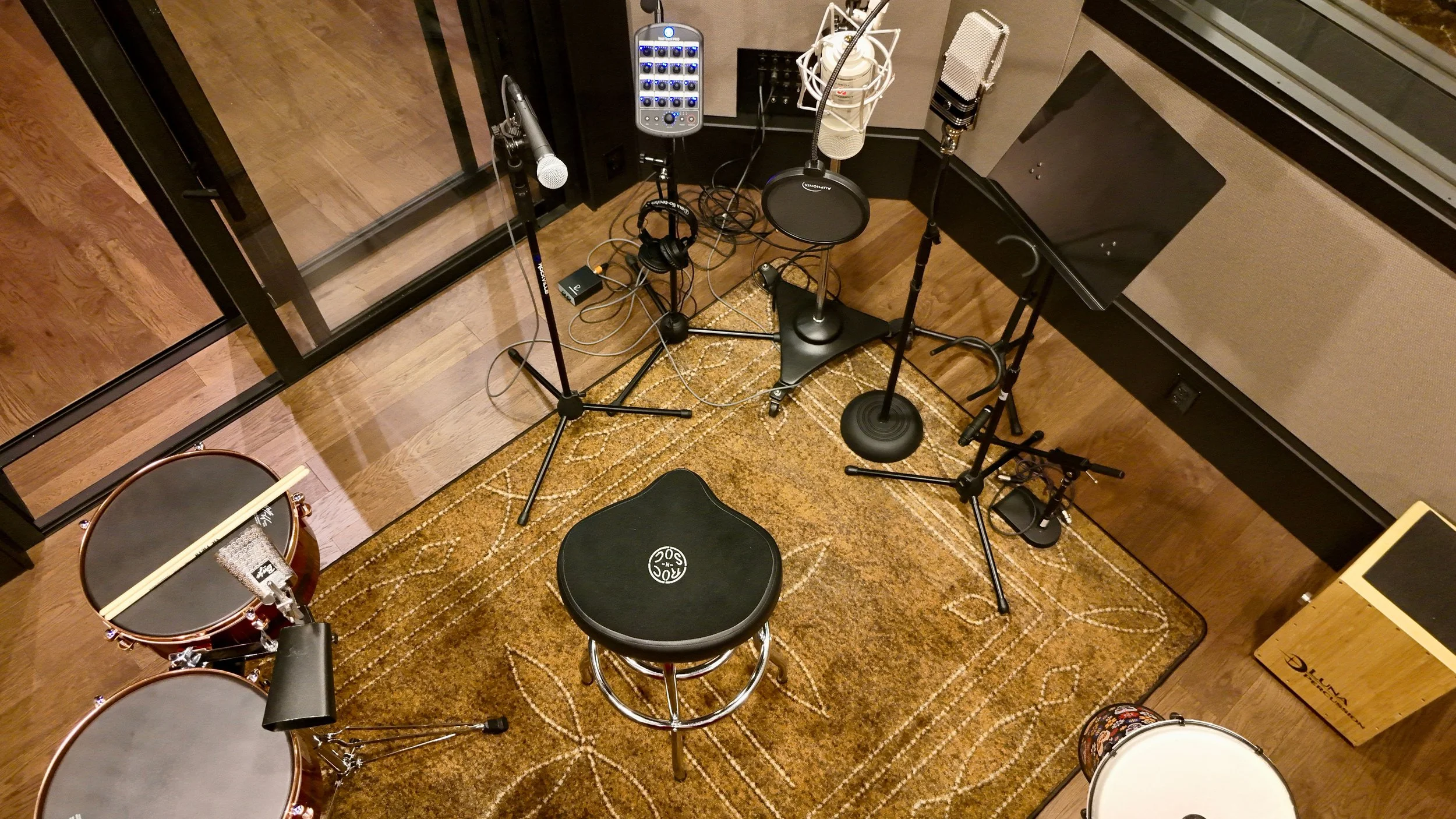 Recording studio setup with microphones, pop filter, headphones, drum set and music stand on a patterned rug.