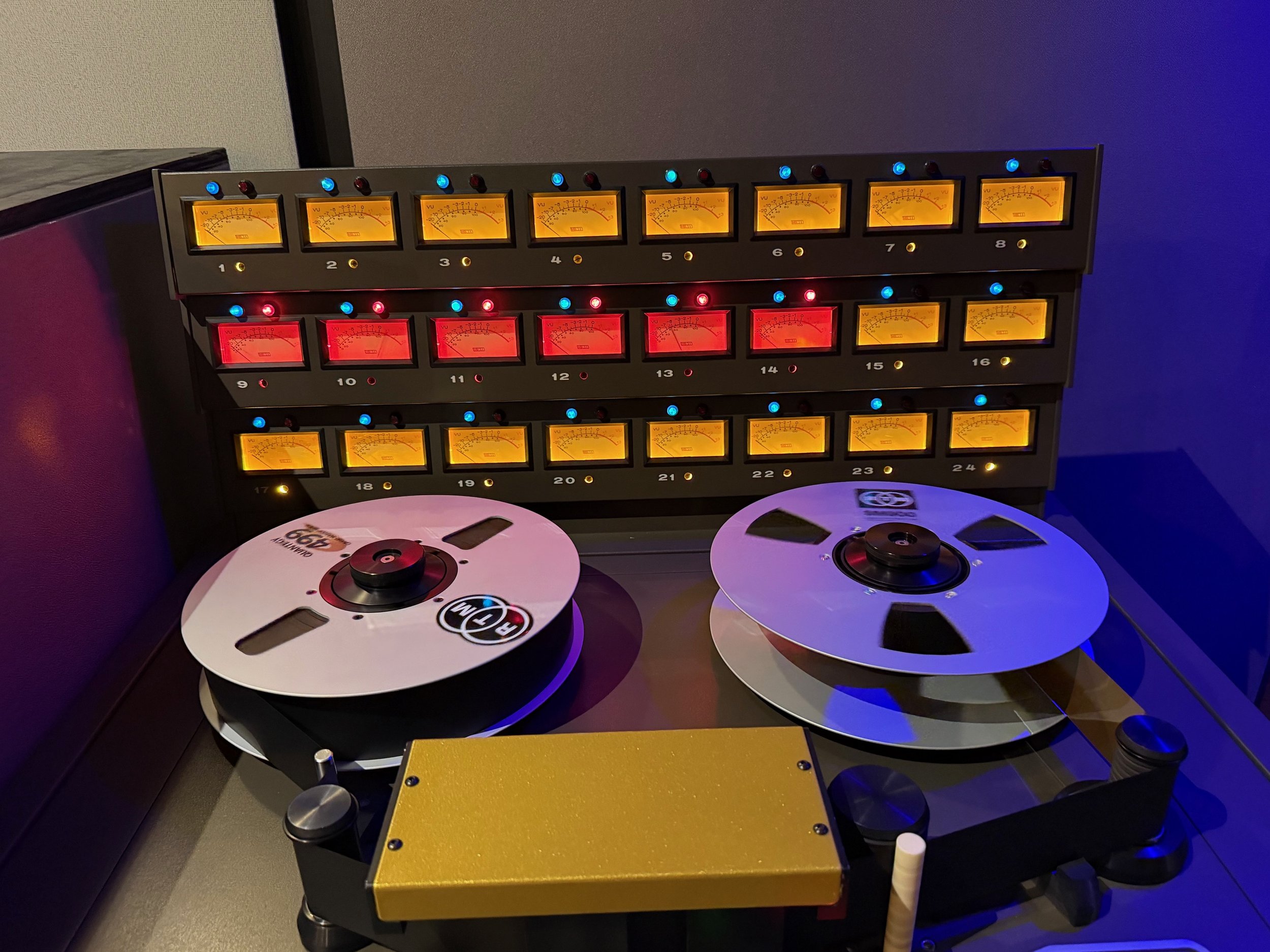 A DJ turntable setup with two turntables, a mixer, and a rack of audio meters with orange and red displays, illuminated by colored lights.