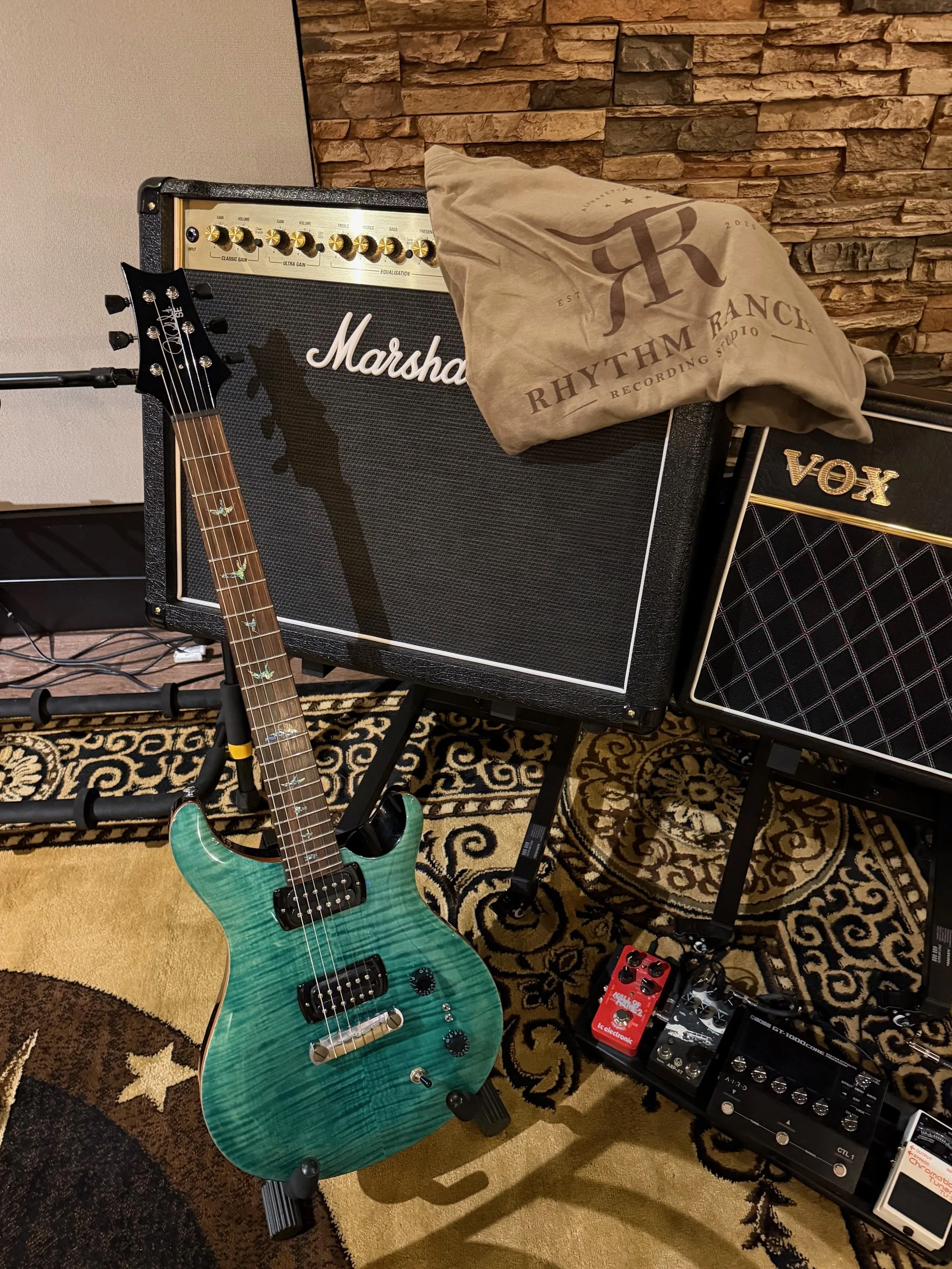 Electric guitar with a green flame top finish leaning against a speaker, with guitar effects pedals on a riser below, and amplifier cabinets behind. The setting appears to be a music studio or rehearsal space with a brick wall and patterned carpet.