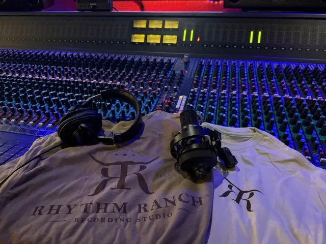 A recording studio mixing console with a pair of headphones and a microphone resting on a T-shirt that reads 'Rhythm Ranch Recording Studio'.