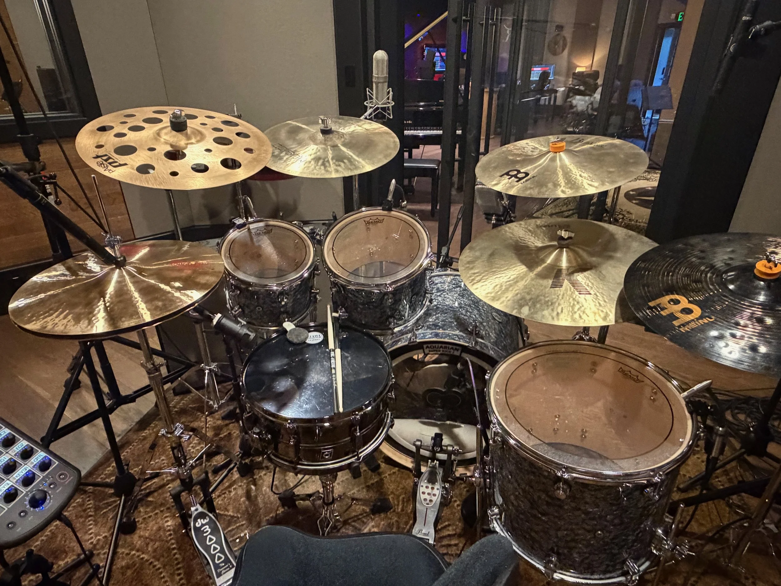 A drum set with multiple cymbals, including hi-hats, crash, and ride cymbals, in a music studio with a microphone nearby, a keyboard in the background, and soundproofing on the walls.