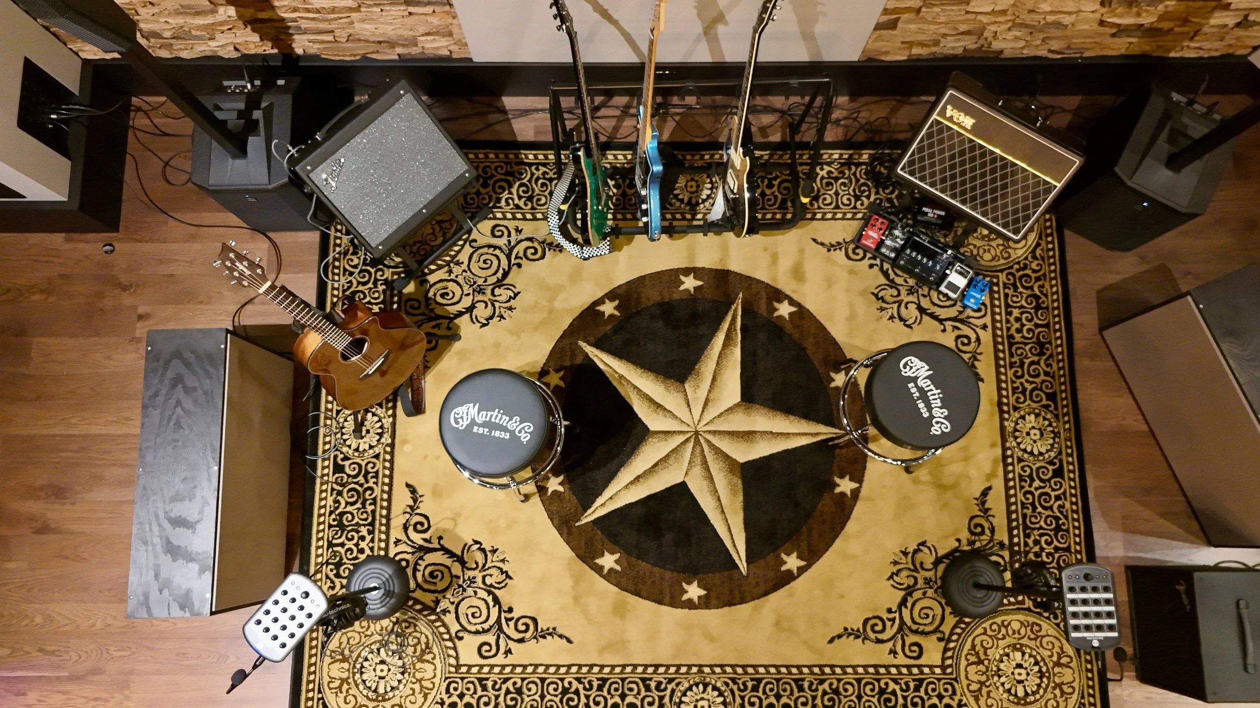 Overview of a music studio or practice space with several guitars on stands, two amplifiers, a small pedalboard, two stools with 'Martin & Co.' written on them, and various audio equipment. The room has a decorative circular rug with a star design, hardwood flooring, and a wall with textured paneling.