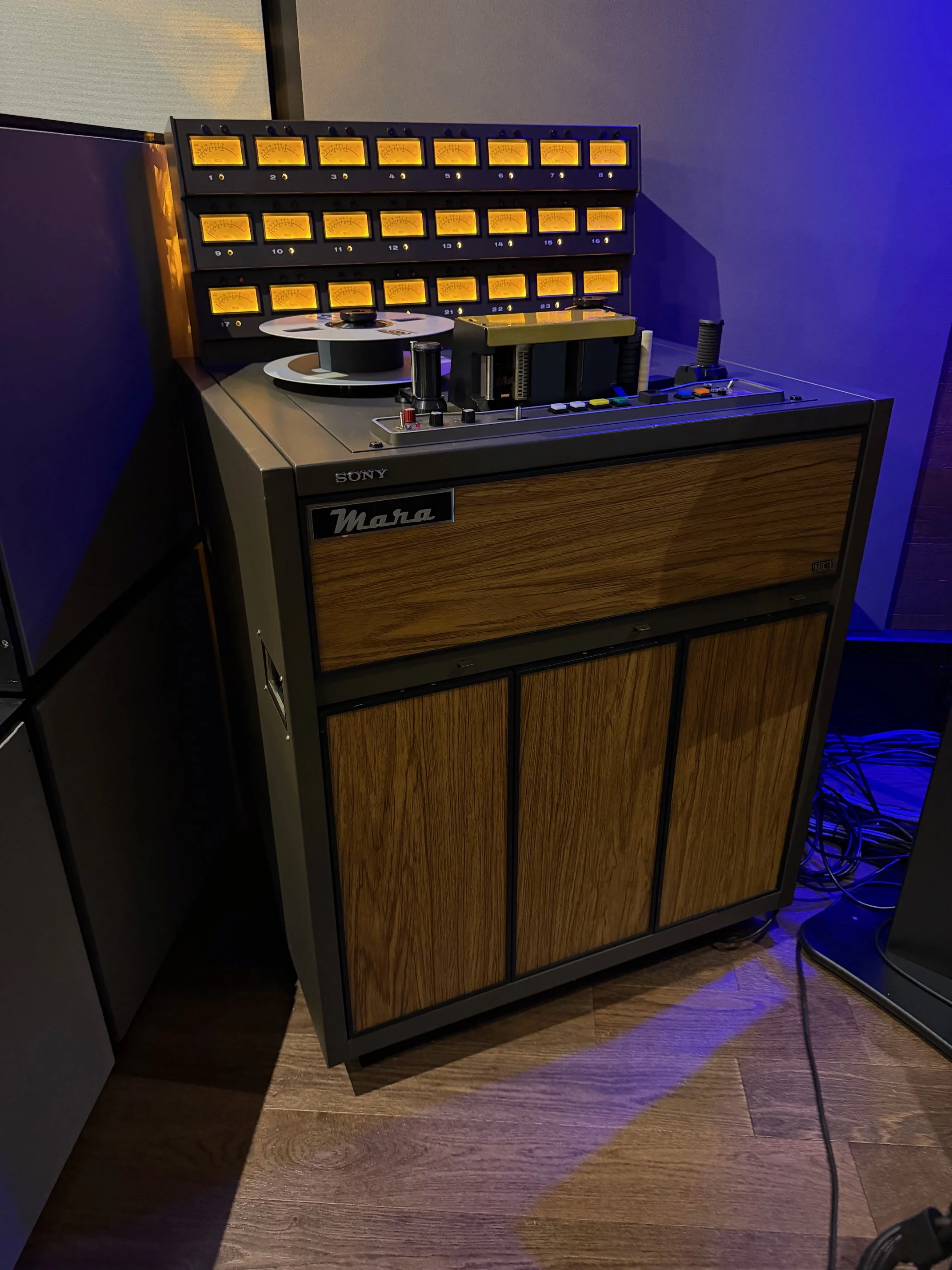 A vintage Sony Mara reel-to-reel tape recorder with a wooden front panel and a black metal frame, placed on a wooden floor, with a blue wall background and a power cable plugged into it.