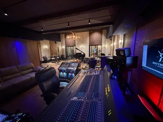 Recording studio with a large mixing console, speakers, and a grand piano in the background, illuminated by warm lighting and a chandelier.