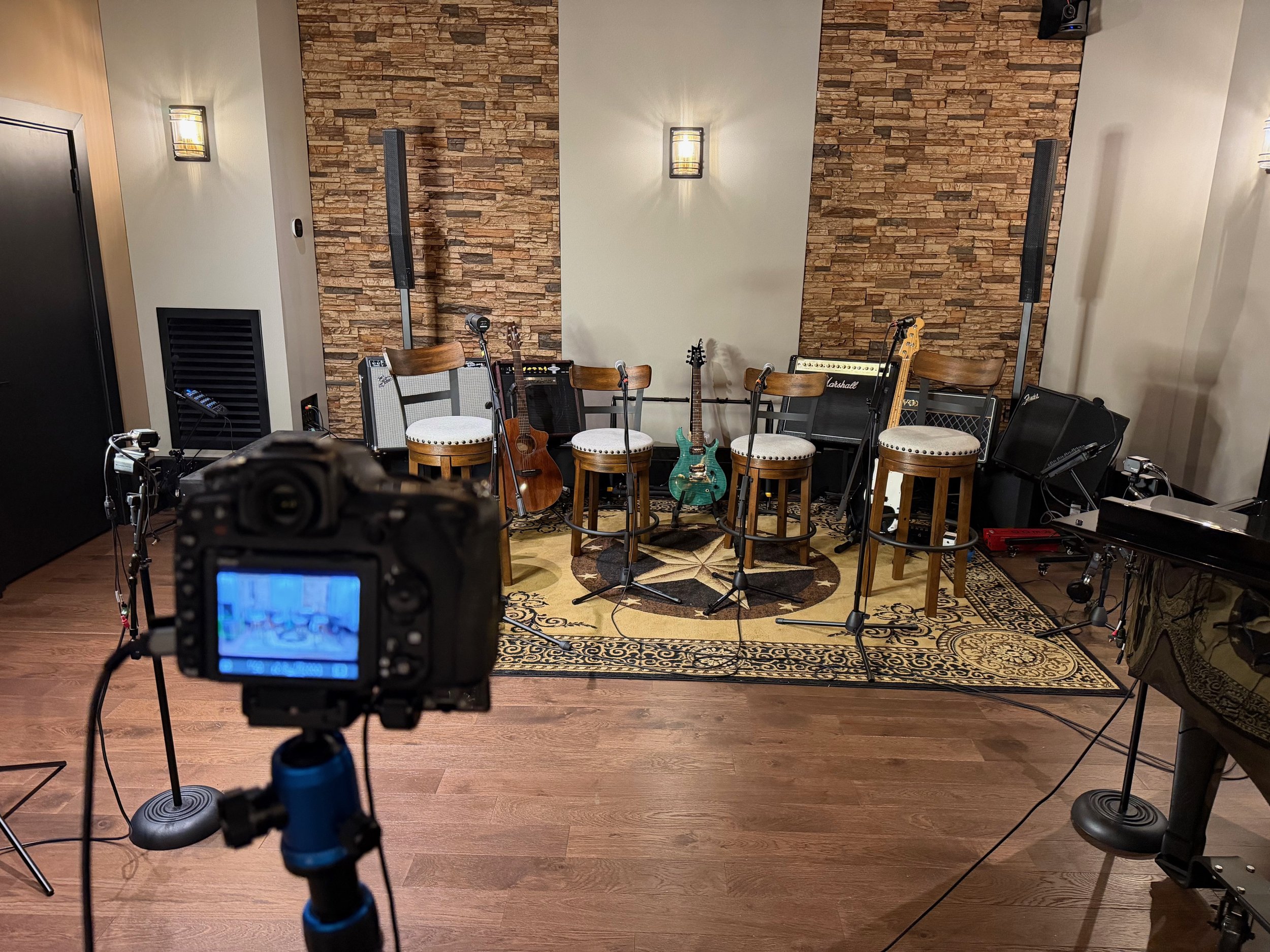 Empty stage with musical instruments including guitars, amplifiers, microphone stands, and chairs, set against a brick wall with two wall-mounted lights and two large speakers.