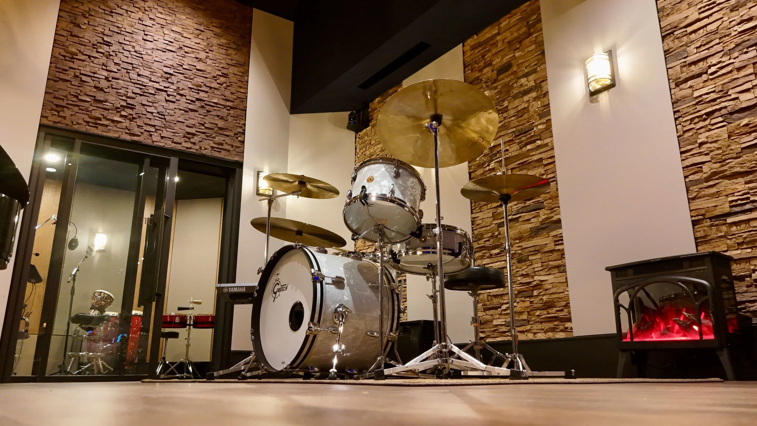 A music practice room with a white Gretsch drum set and cymbals, decorated with a brick accent wall, a fireplace, and some musical equipment.