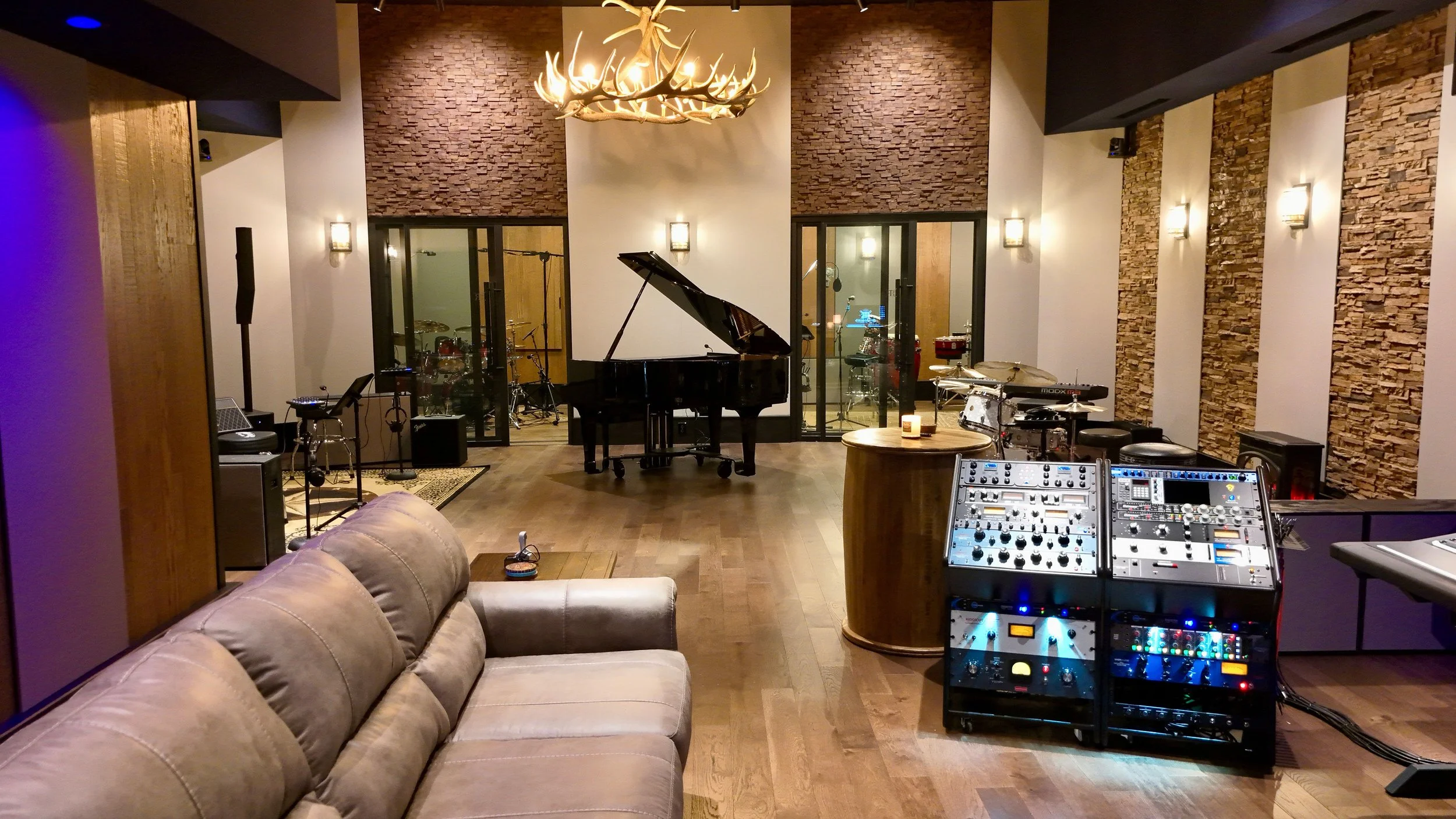 Music studio with a grand piano in the center, surrounded by drums, microphones, and sound equipment, with a cozy sofa in the foreground and a large chandelier overhead.