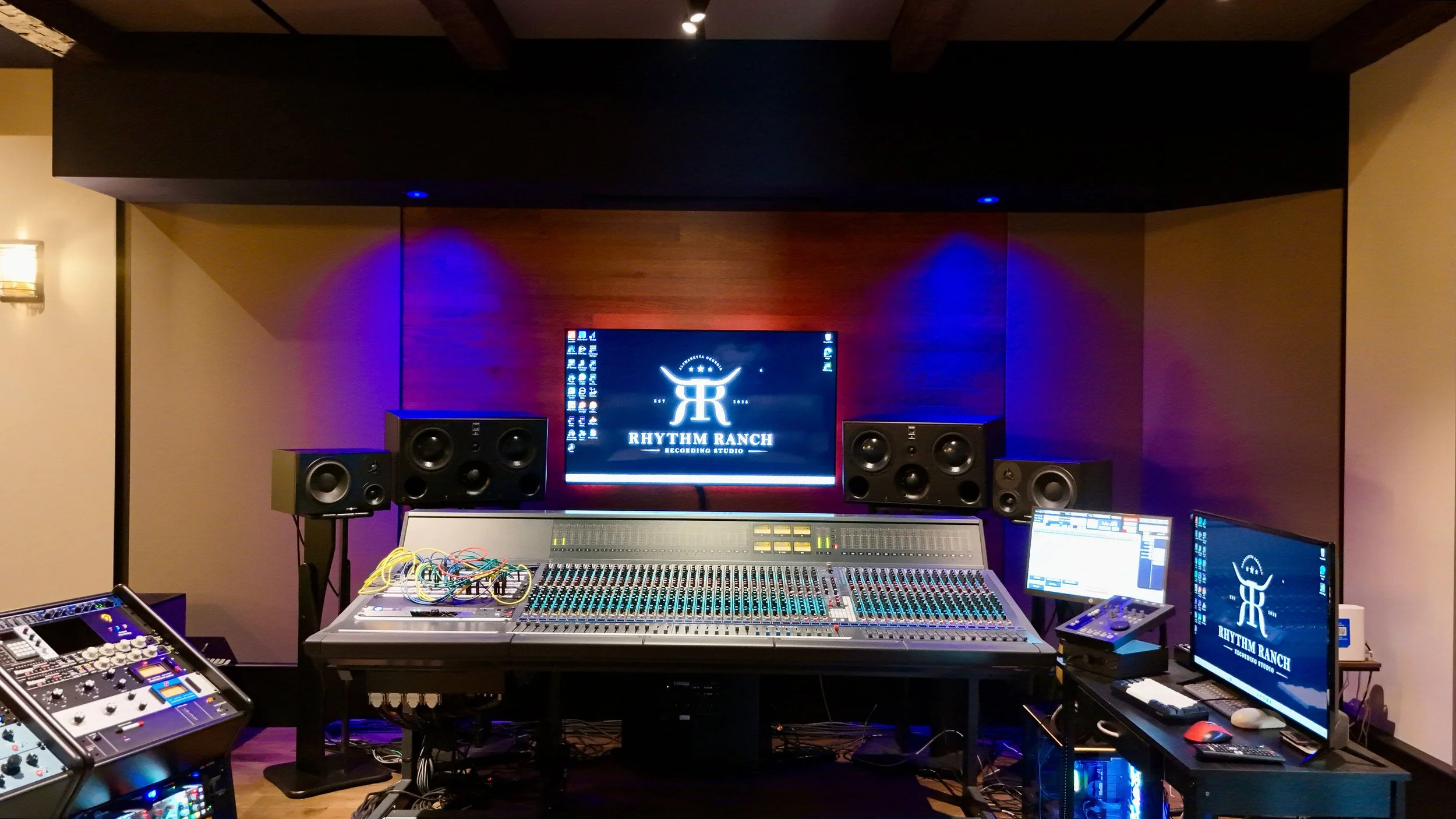 Music recording studio with professional audio equipment, including multiple monitors, speakers, a mixing console, and electronic devices, branded with Rhythmx Ranch.