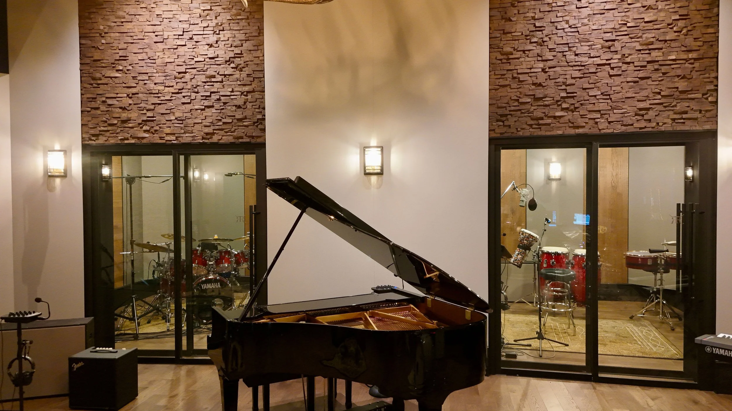 A music studio with a grand piano in the foreground and a drum set visible through glass windows in the background, decorated with wooden acoustic panels and wall-mounted lights.