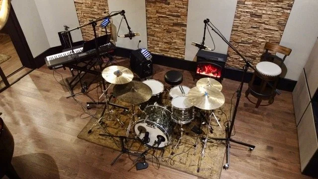Music recording studio with a drum set, keyboard, microphones, and speaker on a wooden floor with stone accent walls.