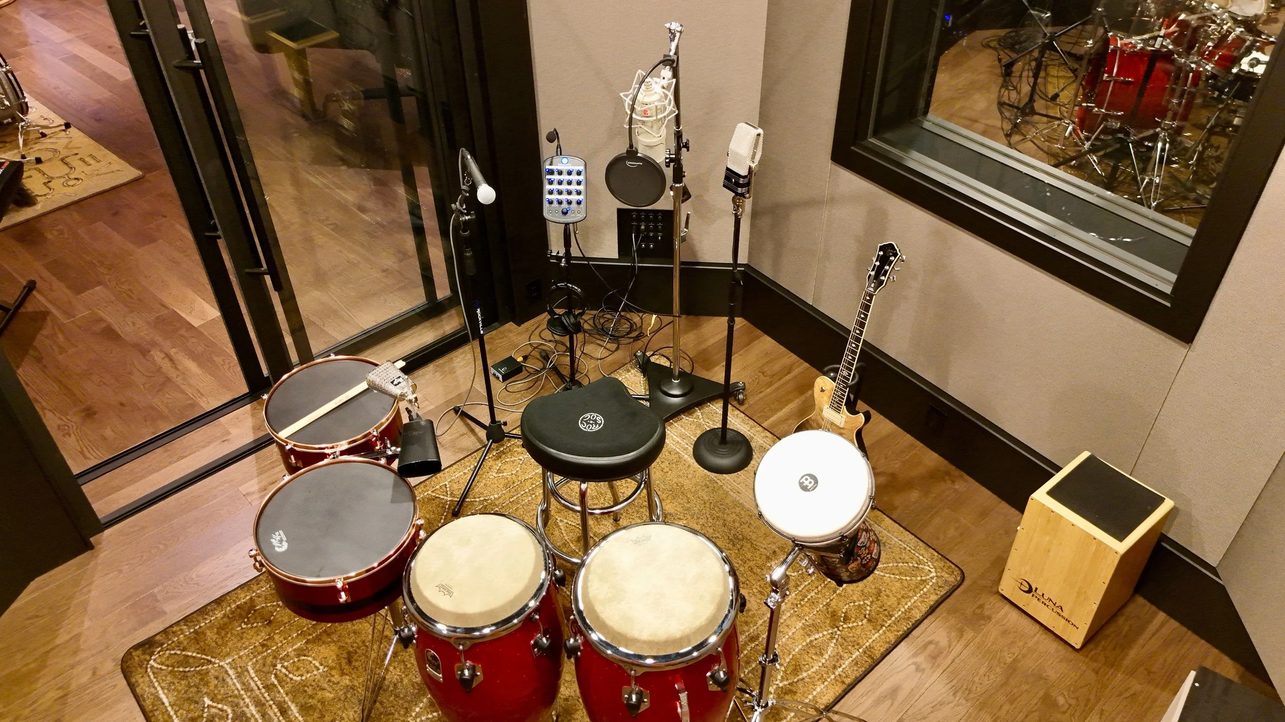 Inside a recording studio with musical instruments, microphones, and audio equipment including drums, a guitar, and a keyboard, viewed through a glass partition.