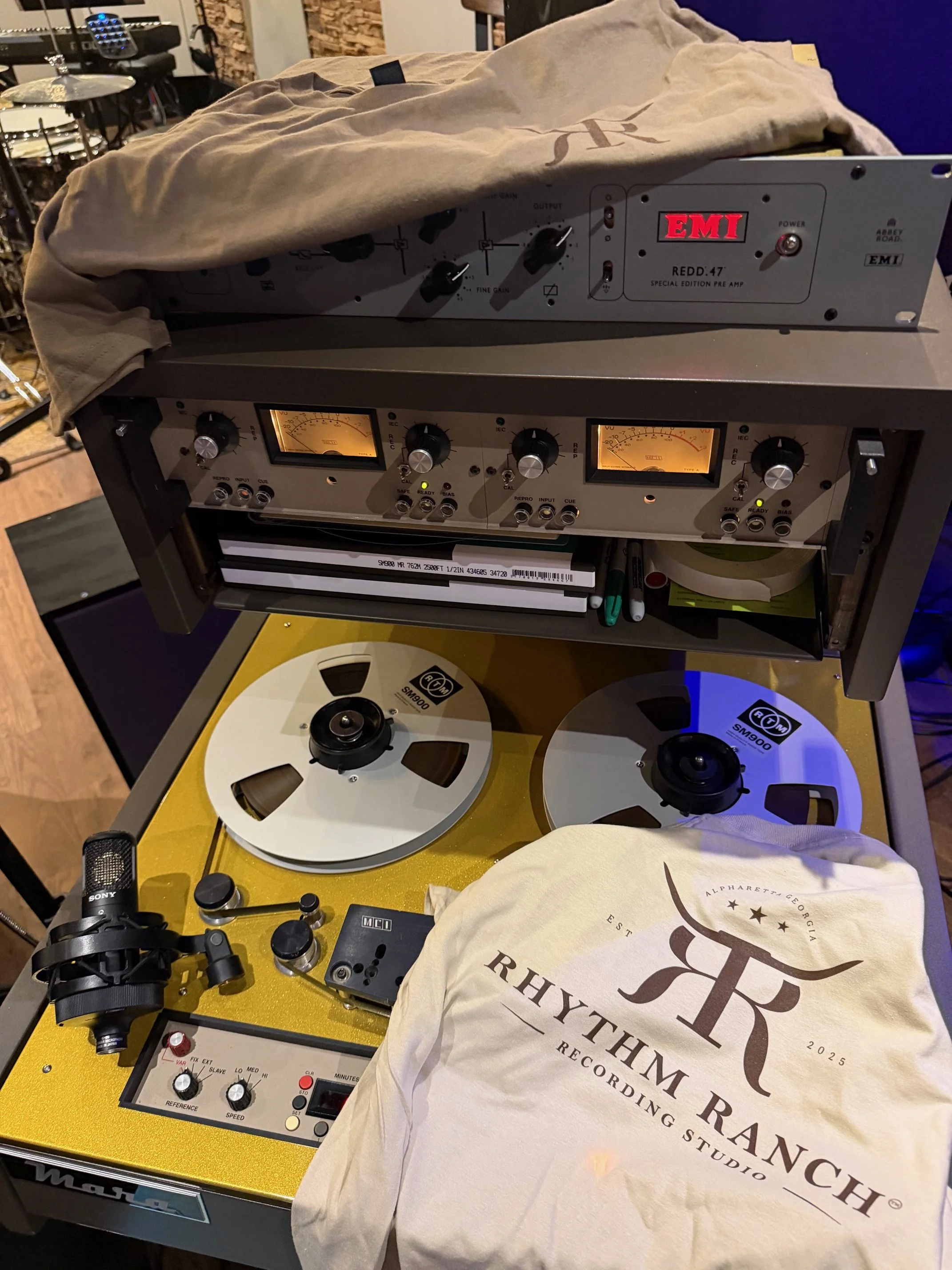 A recording studio setup with a reel-to-reel tape recorder, a control panel with VU meters, and other audio equipment. A cloth bag with the Rhythms Ranch logo is partially covering the equipment, and there are drums and brick walls in the background.