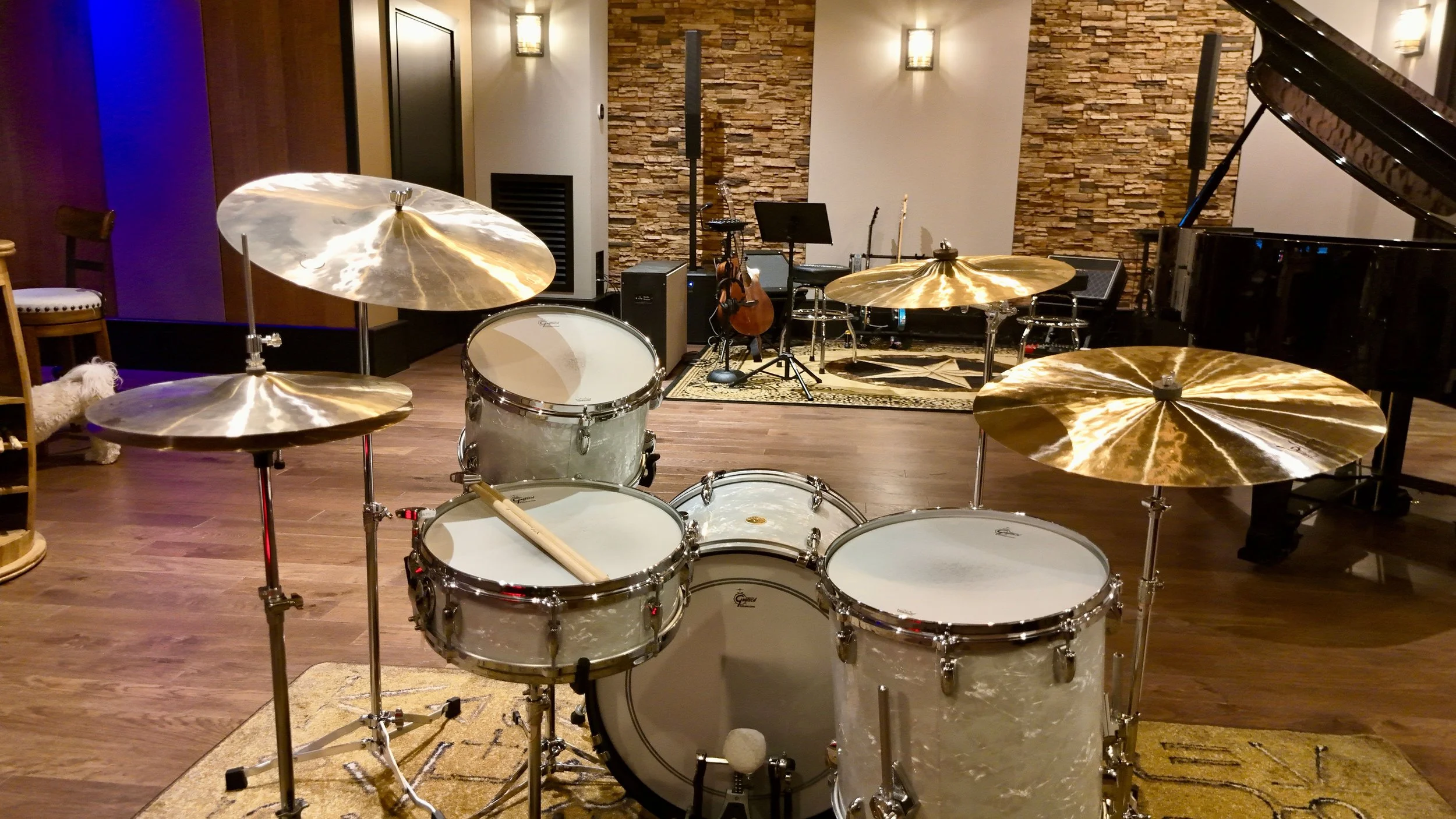 A music studio with a drum set in the foreground, and a grand piano, guitars, and music stands in the background, on a wooden floor with brick and beige walls.