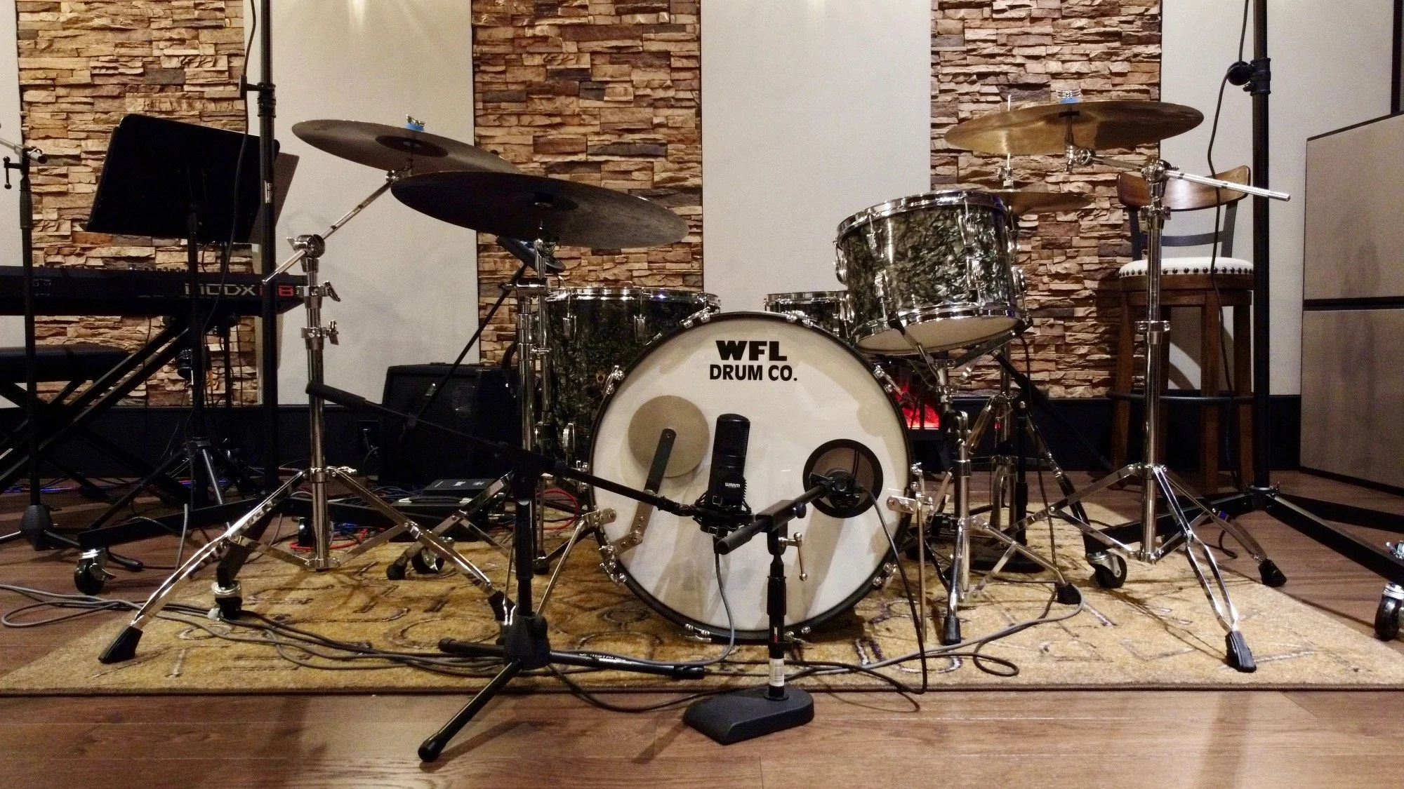 A drum set on stage with a big bass drum labeled 'WFL DRUM CO.', cymbals, drumsticks, and microphones, set against a brick wall backdrop and a small chair on the right.