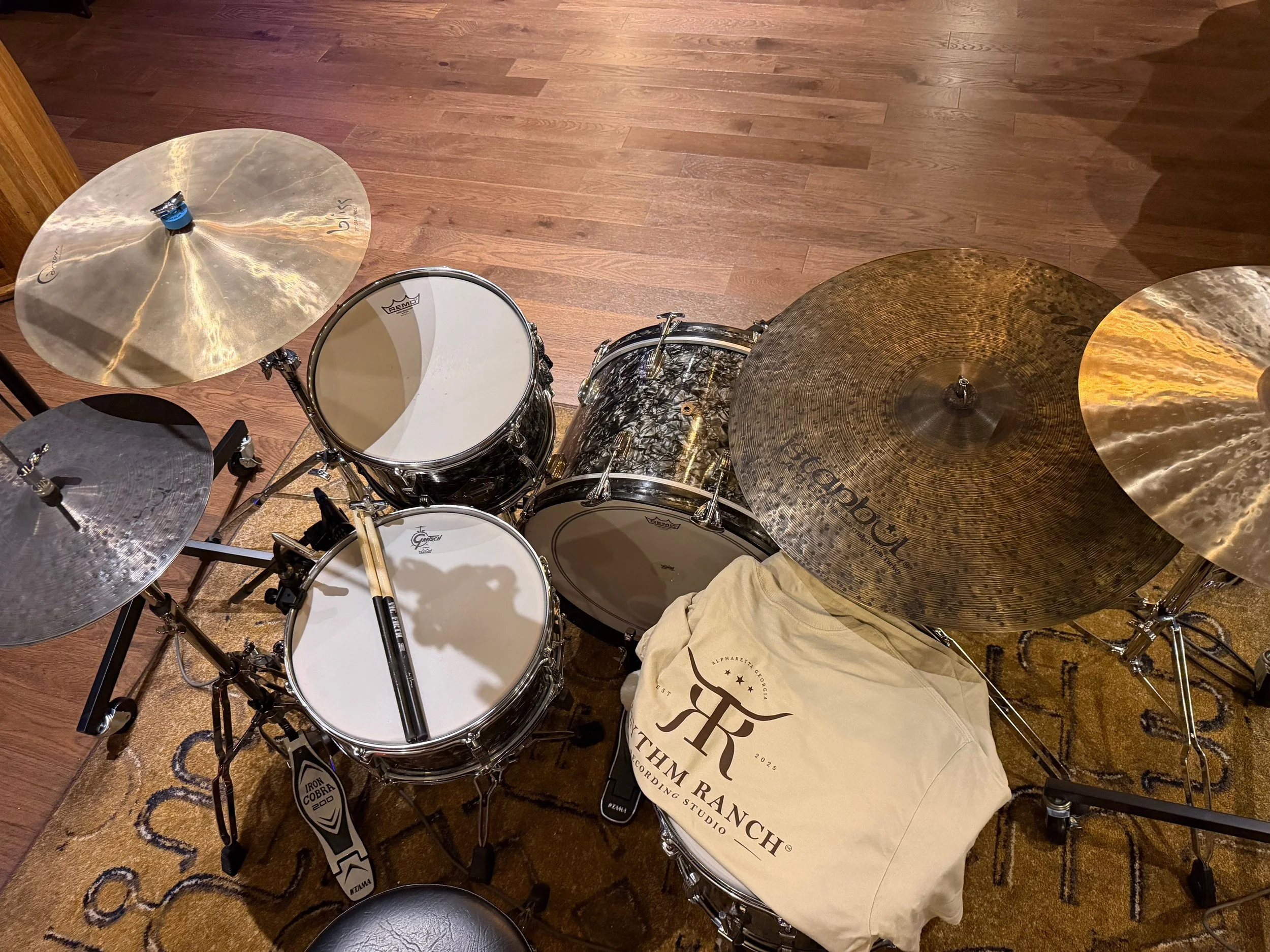 A drum set on a wooden floor with cymbals, a snare drum, and a bass drum, accompanied by a drum throne and a cloth bag with text on it.