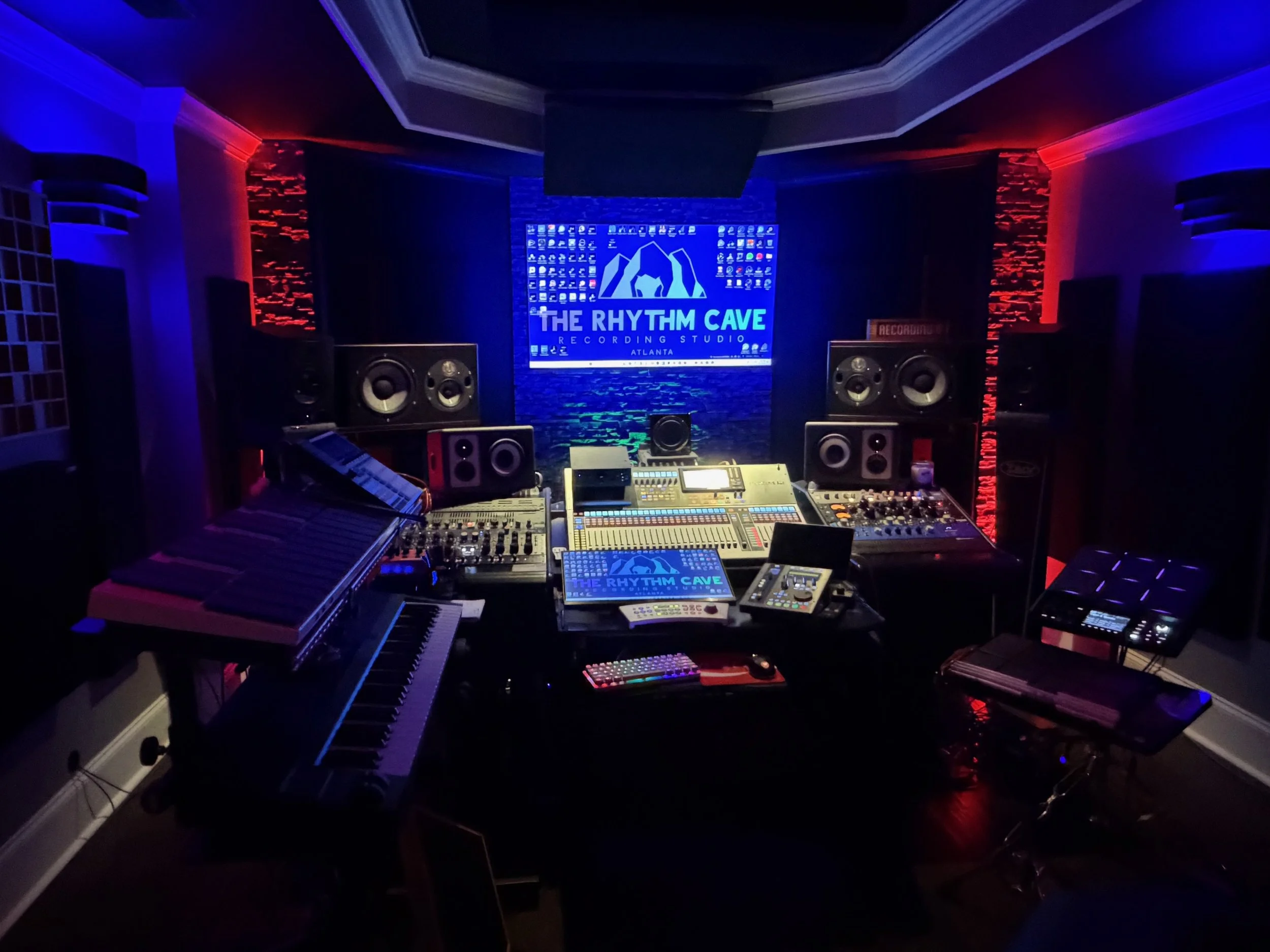 Recording studio with keyboard, mixing console, speakers, and illuminated signage for The Rhythm Cave Recording Studio in Atlanta, featuring colored accent lighting.