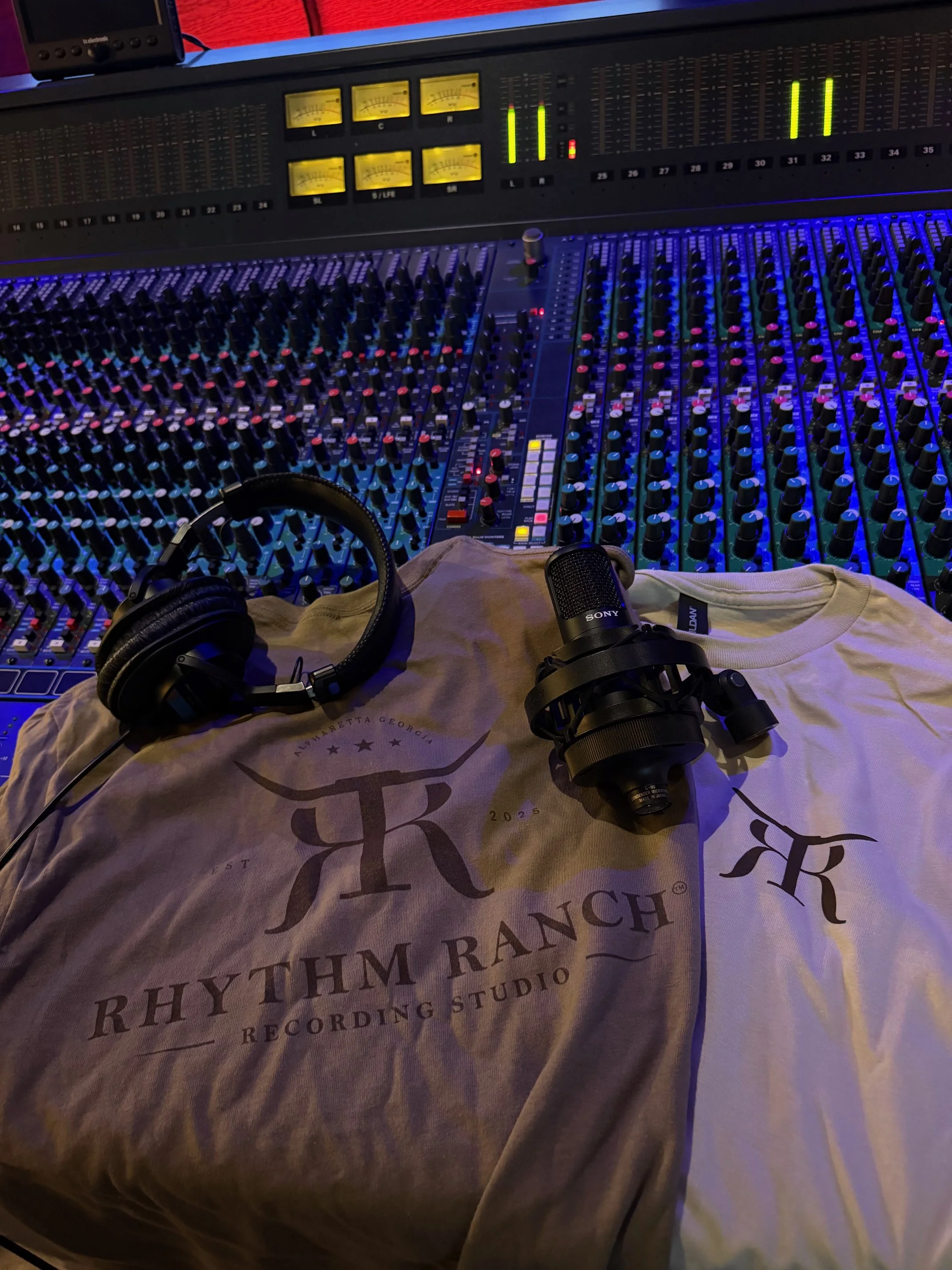 A professional audio mixing console with various controls, meters, headphones, a microphone, and a T-shirt with a logo and text from Rhythim Rhythms Recording Studio.