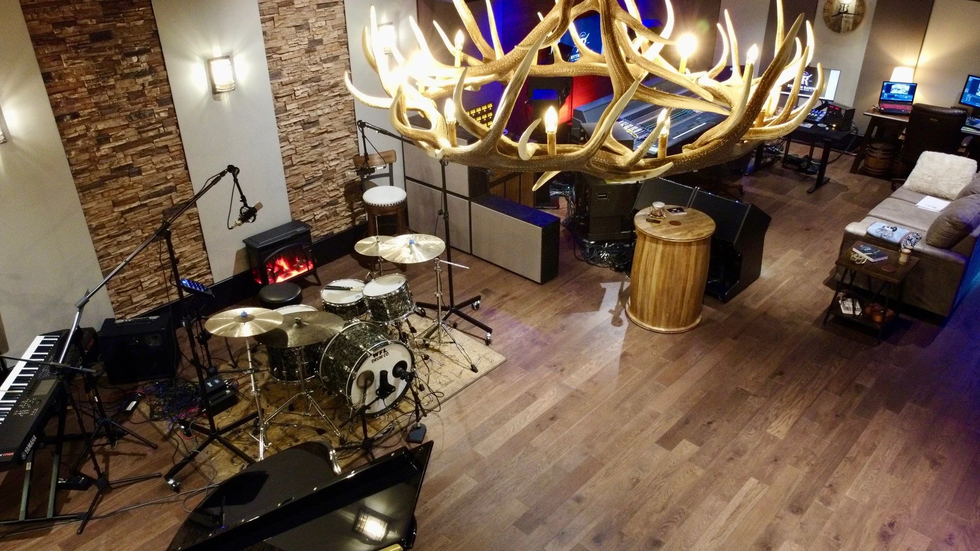 A cozy, well-lit music studio with a large chandelier made of antlers hanging from the ceiling. The room features a drum set with a bass drum, cymbals, and drums, a keyboard, and multiple laptops and electronic equipment on tables. There is a sofa with a pillow, a side table with coffee cups, and a textured brick wall on one side with wall-mounted lamps, creating a warm atmosphere for musical practice or recording.