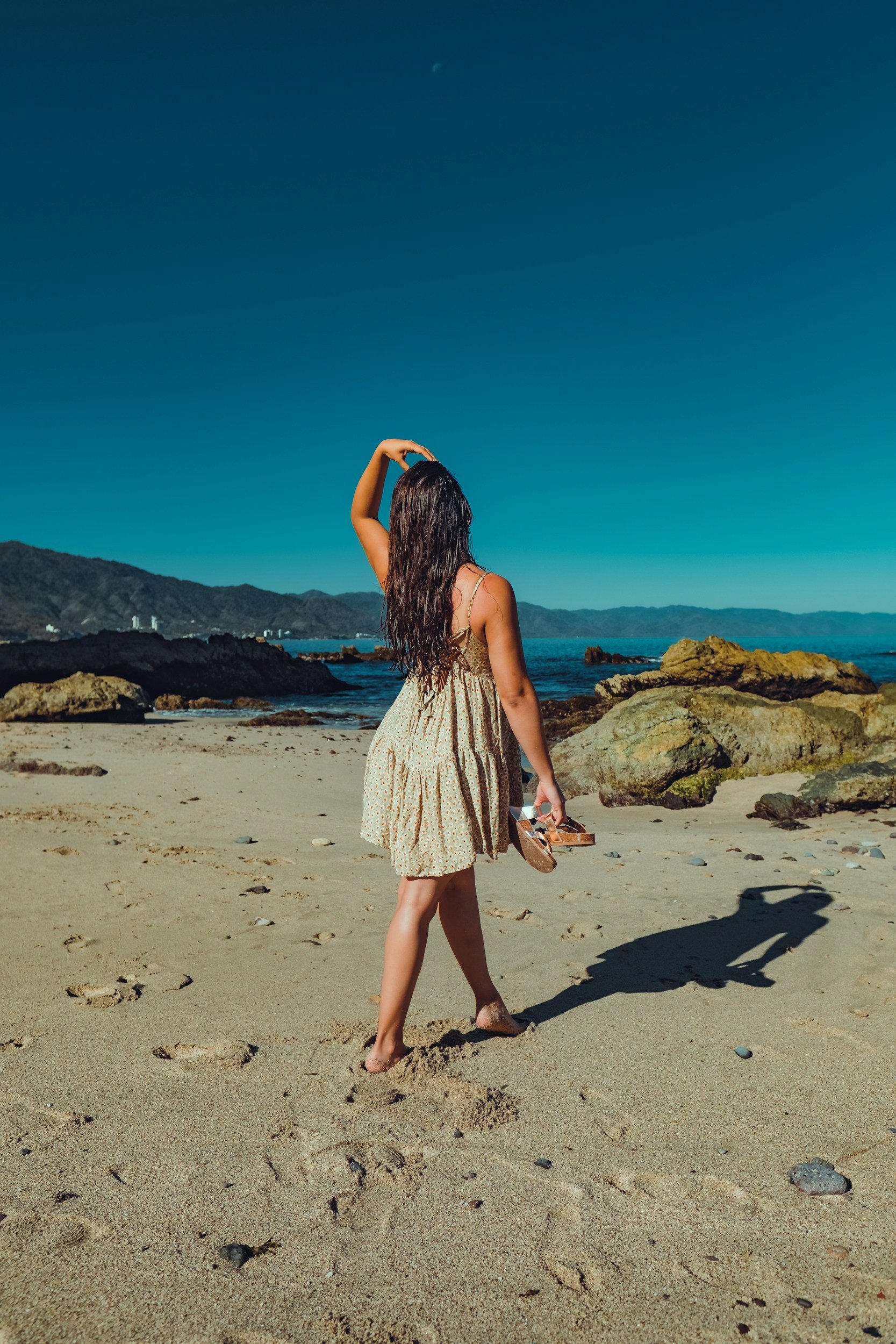 A woman with long dark hair wearing a yellow floral dress walking on a sandy beach, holding a pair of sandals, with rocks and the ocean in the background under a clear blue sky.