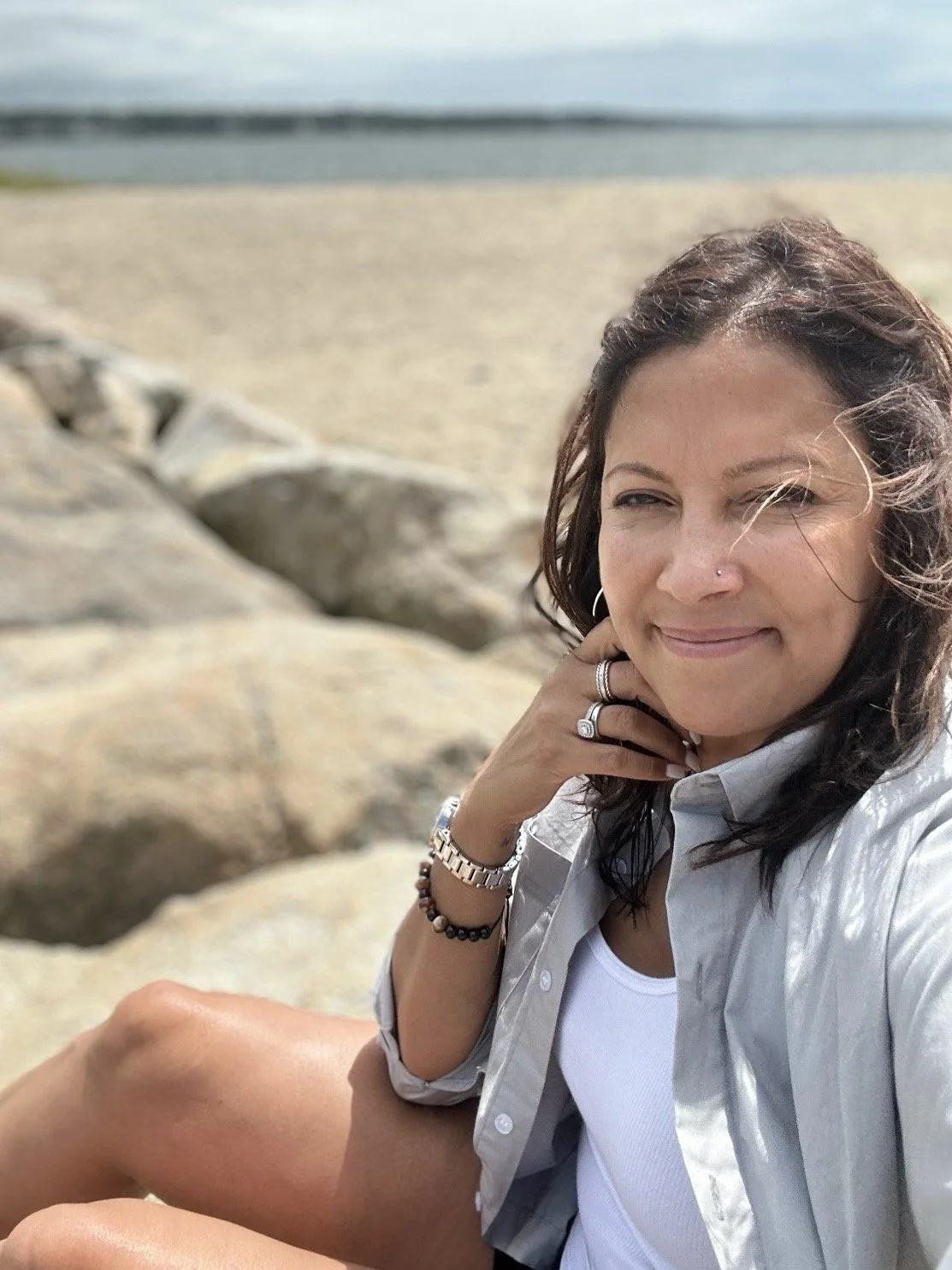 A woman sitting on rocks at the beach, wearing a white shirt and smiling.