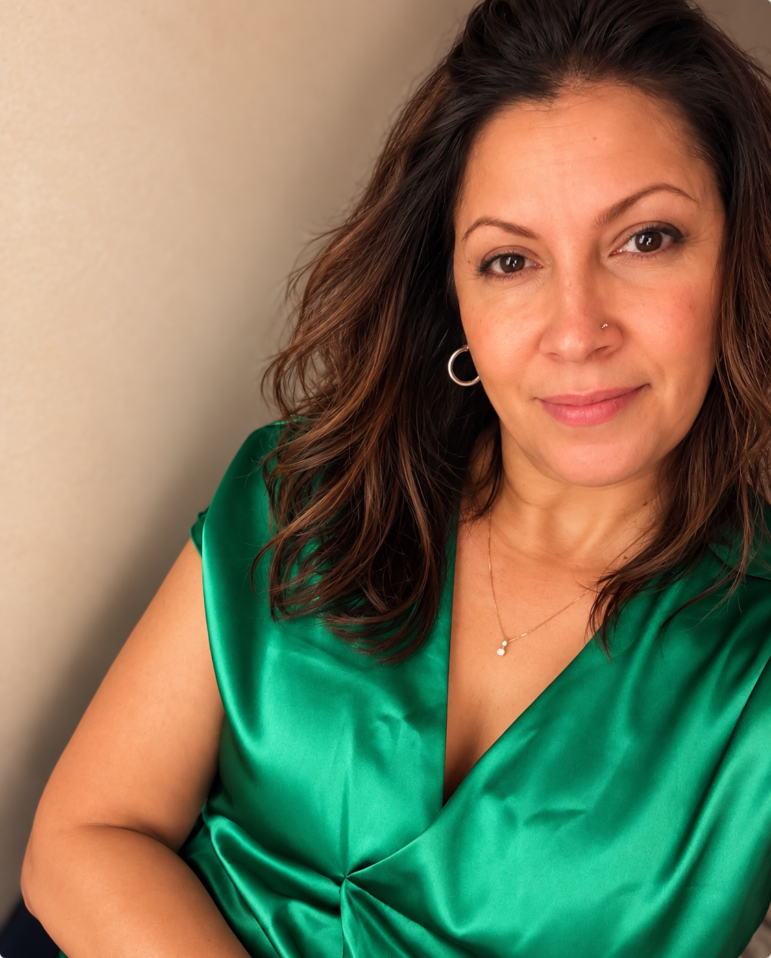 A woman in a emerald green satin top smiling softly against a beige wall.