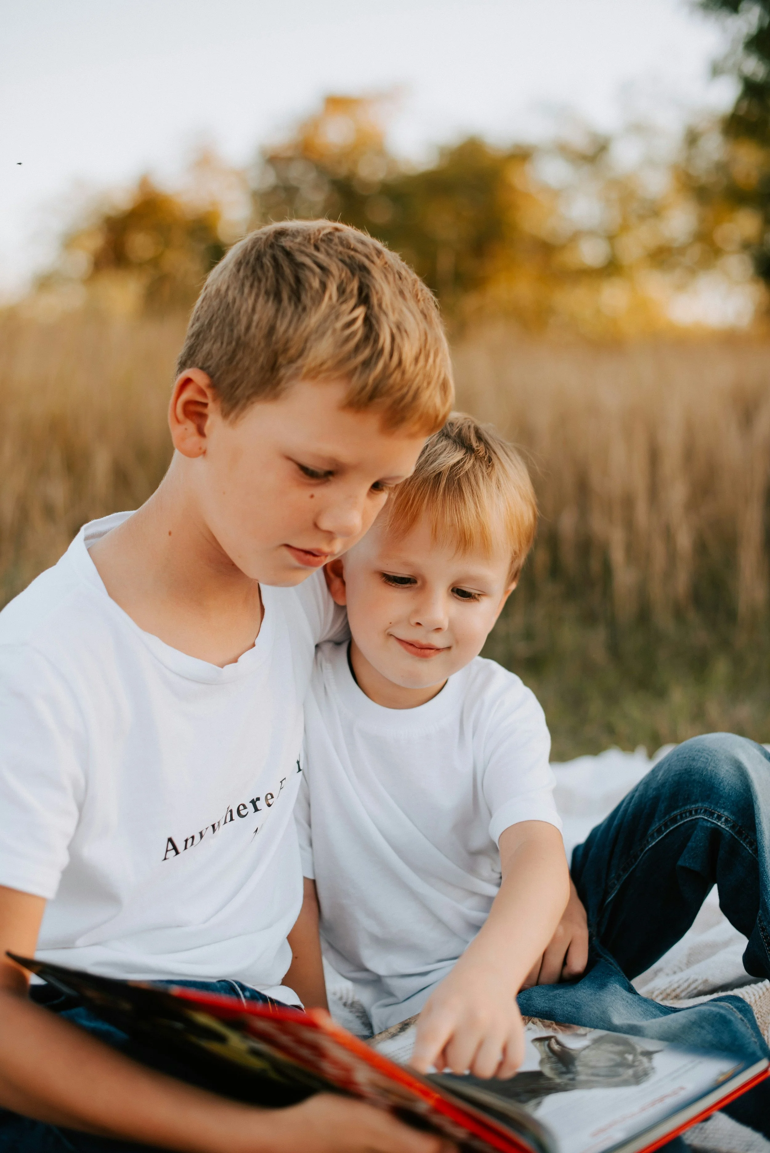 Two young boys with light brown hair and wearing white t-shirts sitting outdoors on a blanket, looking at a photo album together during daytime, with a background of blurred trees and sky.
