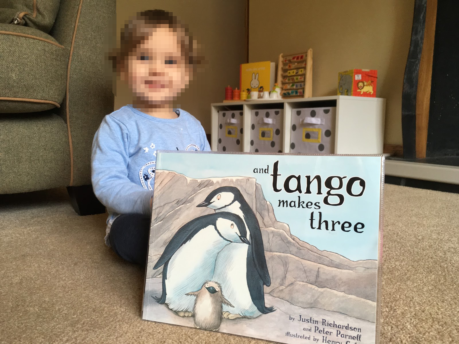 A young child sitting on the carpeted floor holding a children's book titled "and tango makes three" with illustrations of two penguins and a chick on the cover.