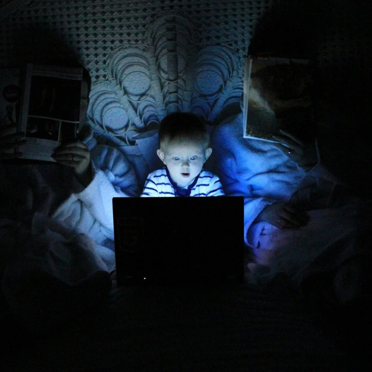 A young boy sitting on a bed in a dark room, illuminated by the glow of a laptop screen, looking surprised or excited.
