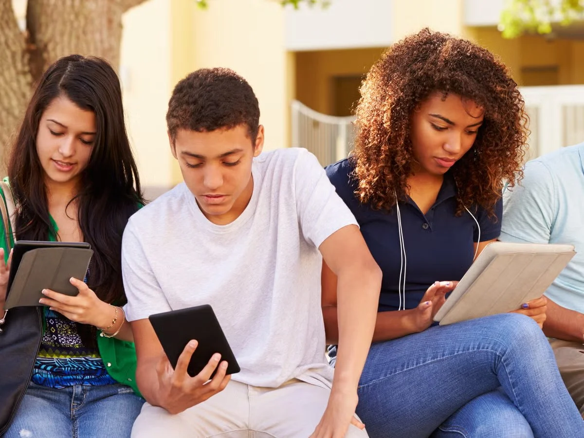 Three teenagers sitting outdoors, engaged with their electronic devices, with trees and a building in the background.