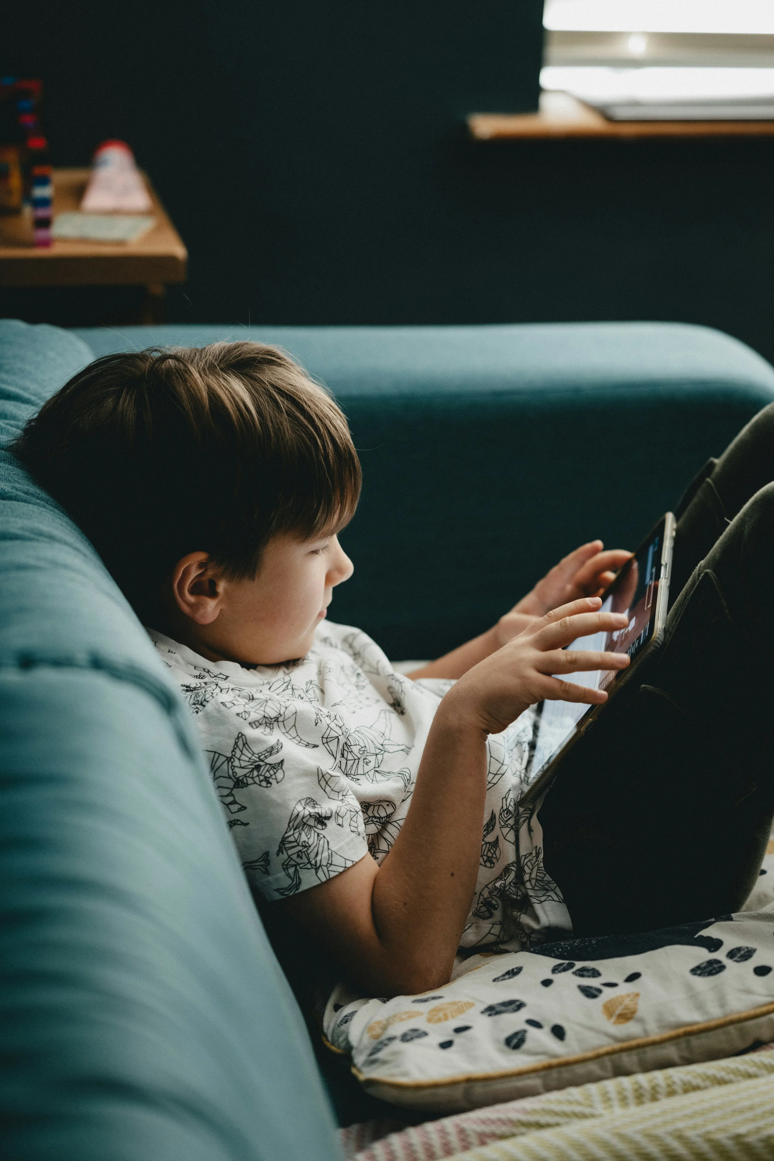 A young boy with brown hair lying on a teal couch, looking at a tablet device, wearing a white T-shirt with black animal sketches.