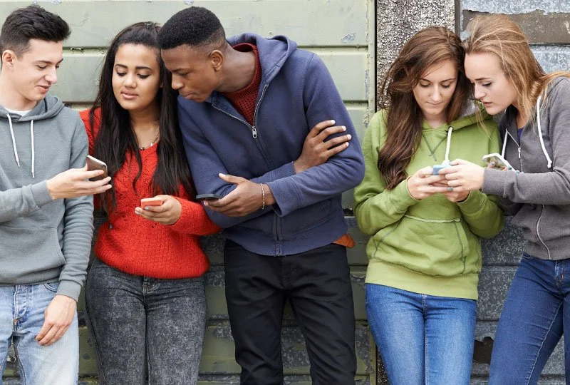Four teenagers standing outdoors against a wooden and concrete wall, all looking at their smartphones.