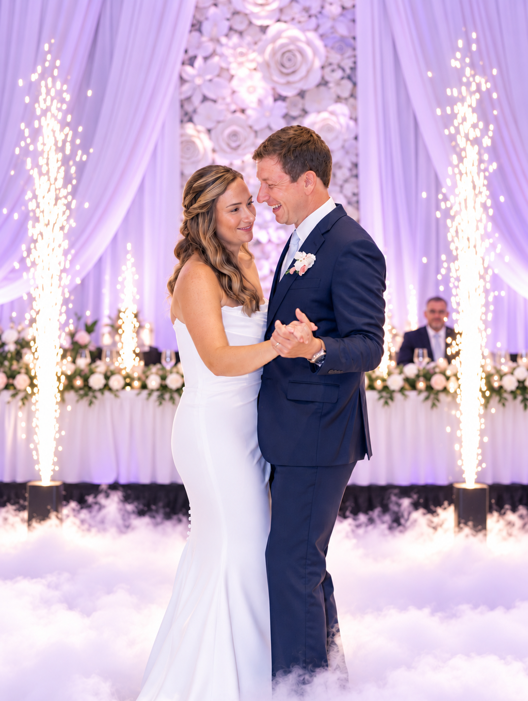 Bride and groom sharing their first dance on a low cloud of dry ice in front of a flower wall and purple uplighting