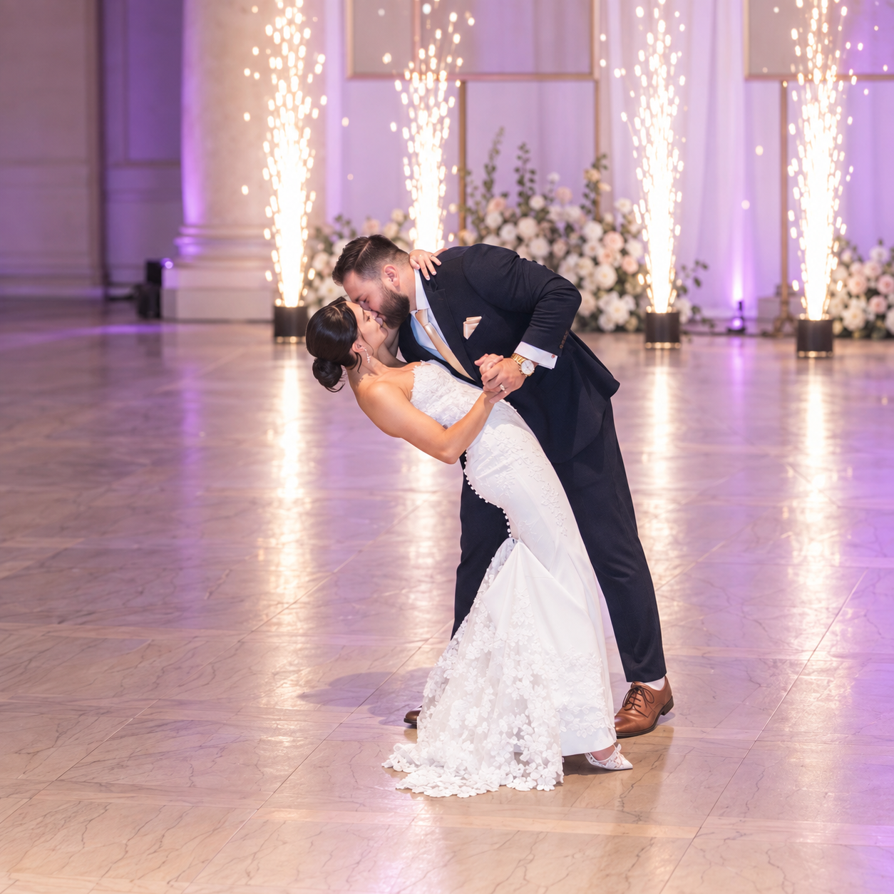 Groom dipping the bride for a kiss on the dance floor with cold spark fountains and purple uplighting behind them