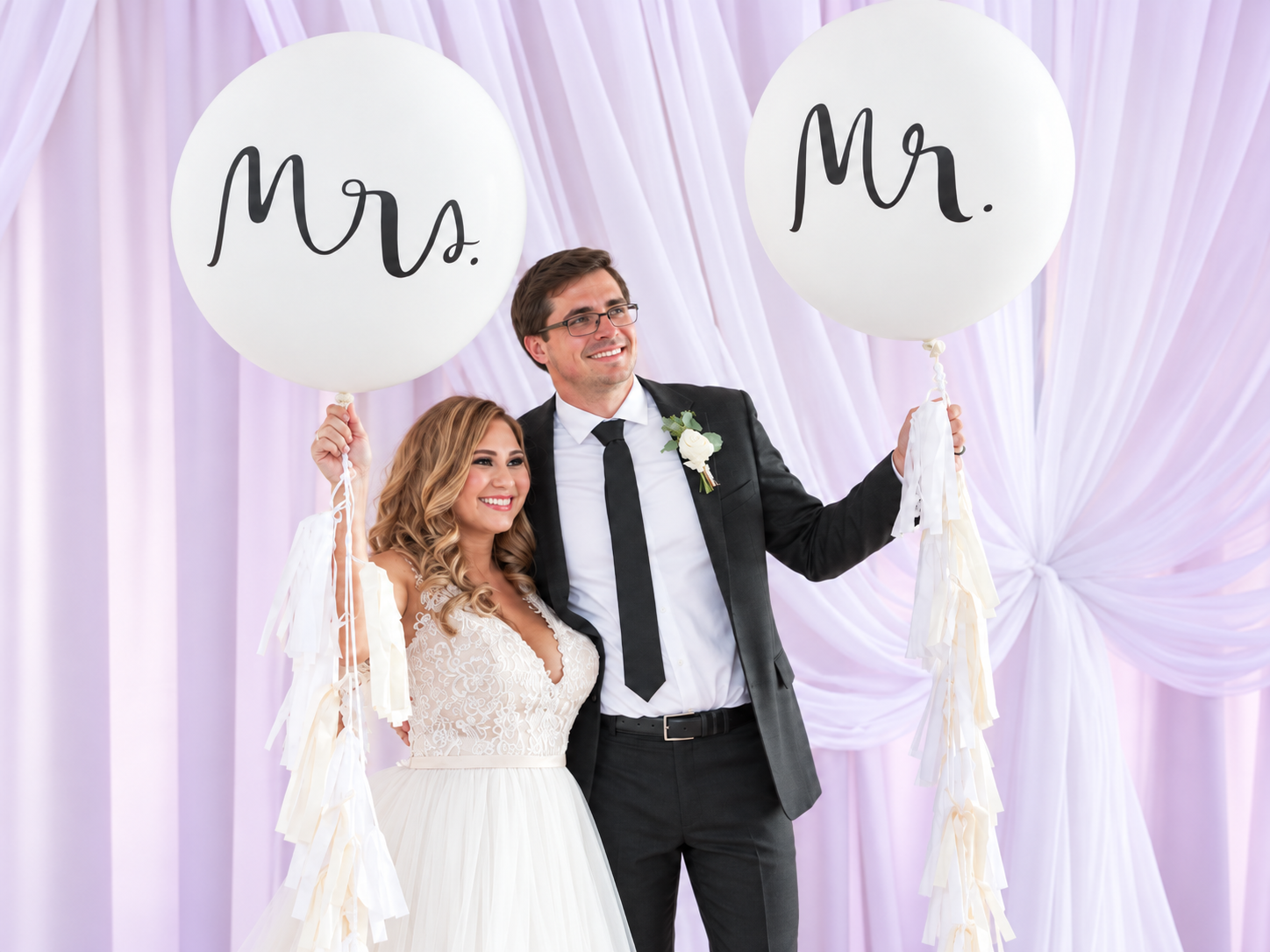 Smiling bride and groom holding giant Mr. and Mrs. balloons in front of draped fabric