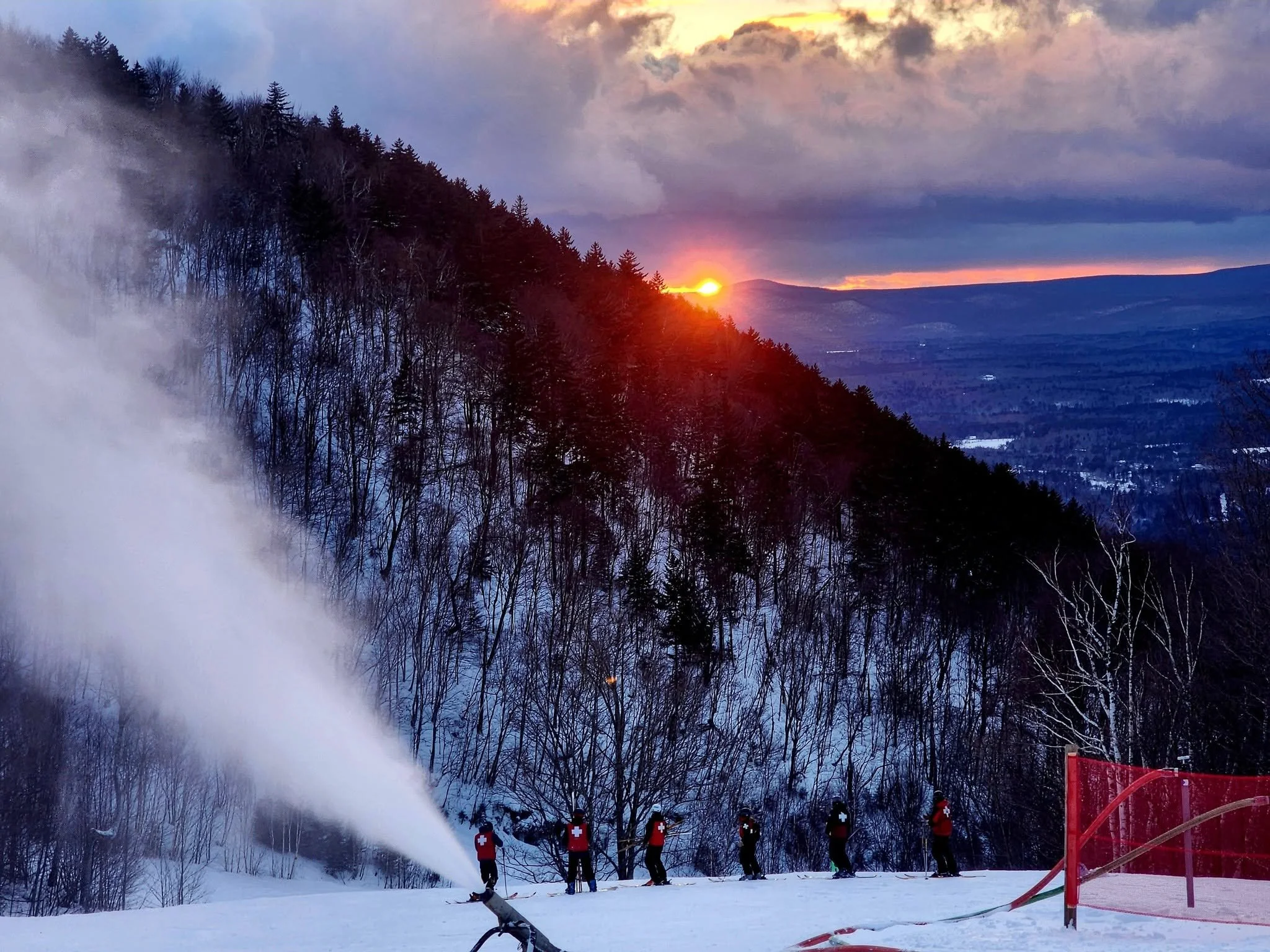 Skiers with red jackets and white crosses on their backs stand in line on snow-covered ground, with a snow cannon spraying snow nearby. The scene is set in a mountainous area with leafless trees, and a sunset or sunrise in the sky with clouds and a hint of sunlight.