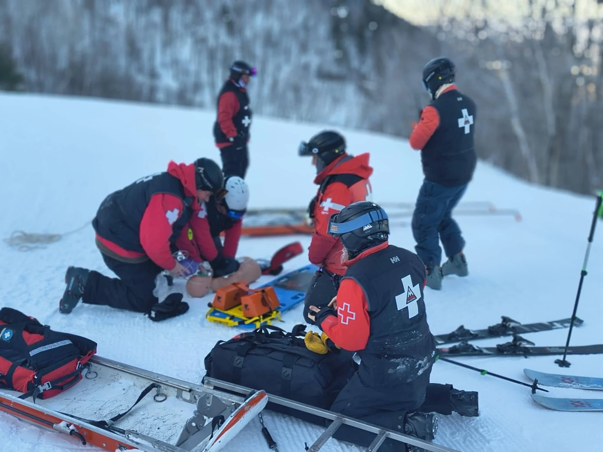 A group of rescue workers in winter gear attending to an injured person lying on a stretcher in a snow-covered outdoor area, with rescue equipment around them.