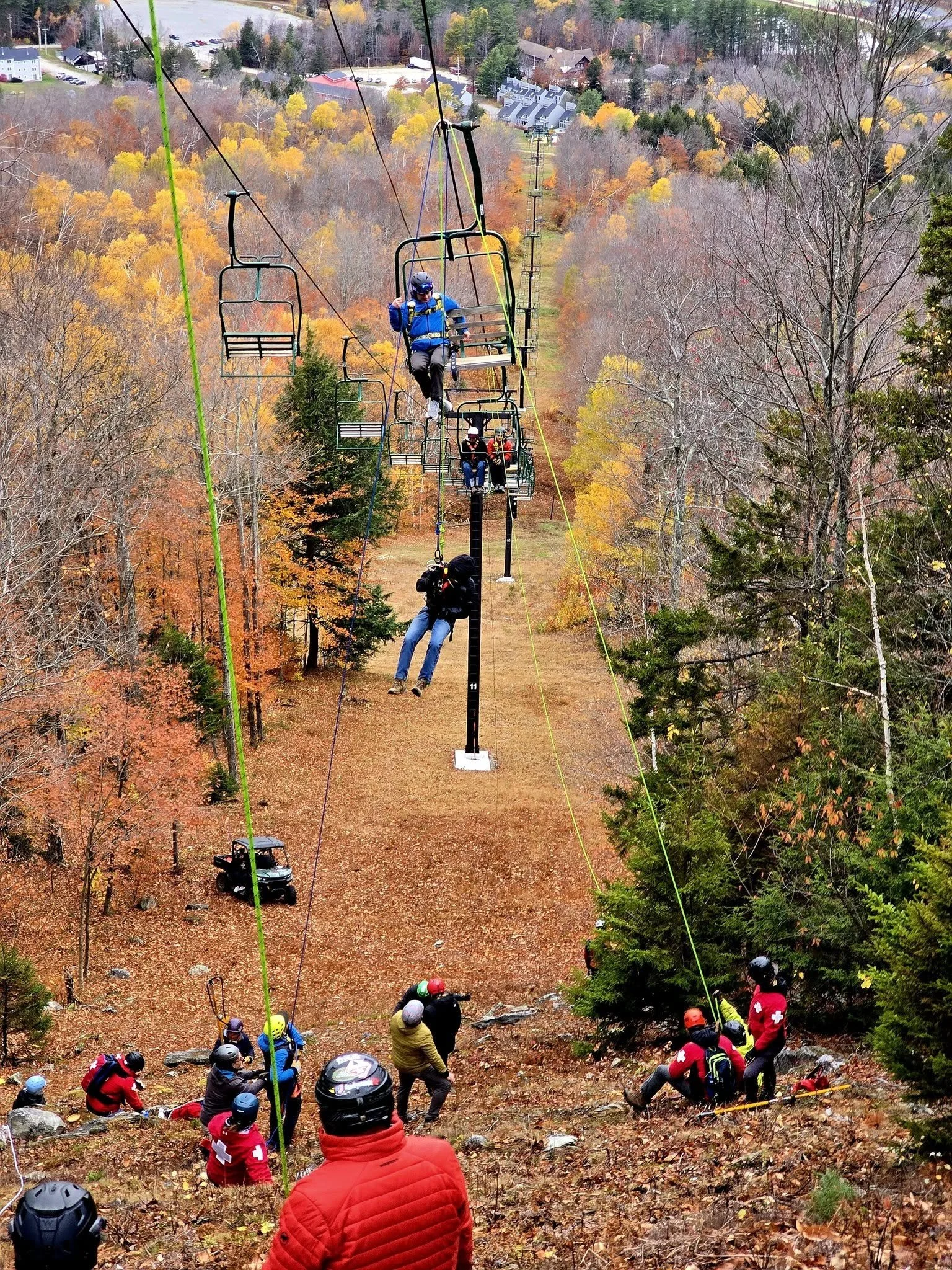 A group of people dressed in winter gear and helmets waiting on a hillside covered in fallen leaves, with a ski lift passing overhead and a forest with autumn foliage in the background.