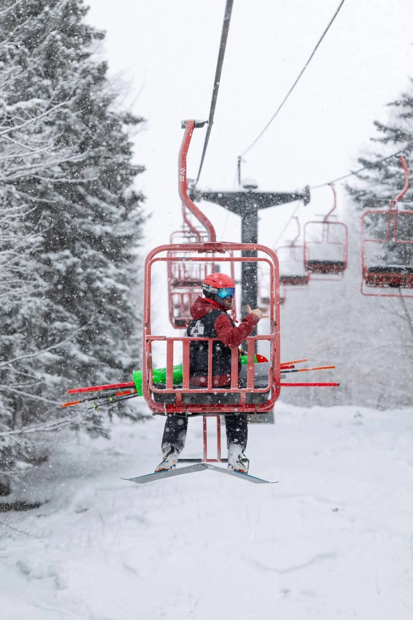 A person wearing a red helmet and ski gear riding a ski lift through a snowy winter landscape lined with snow-covered trees.