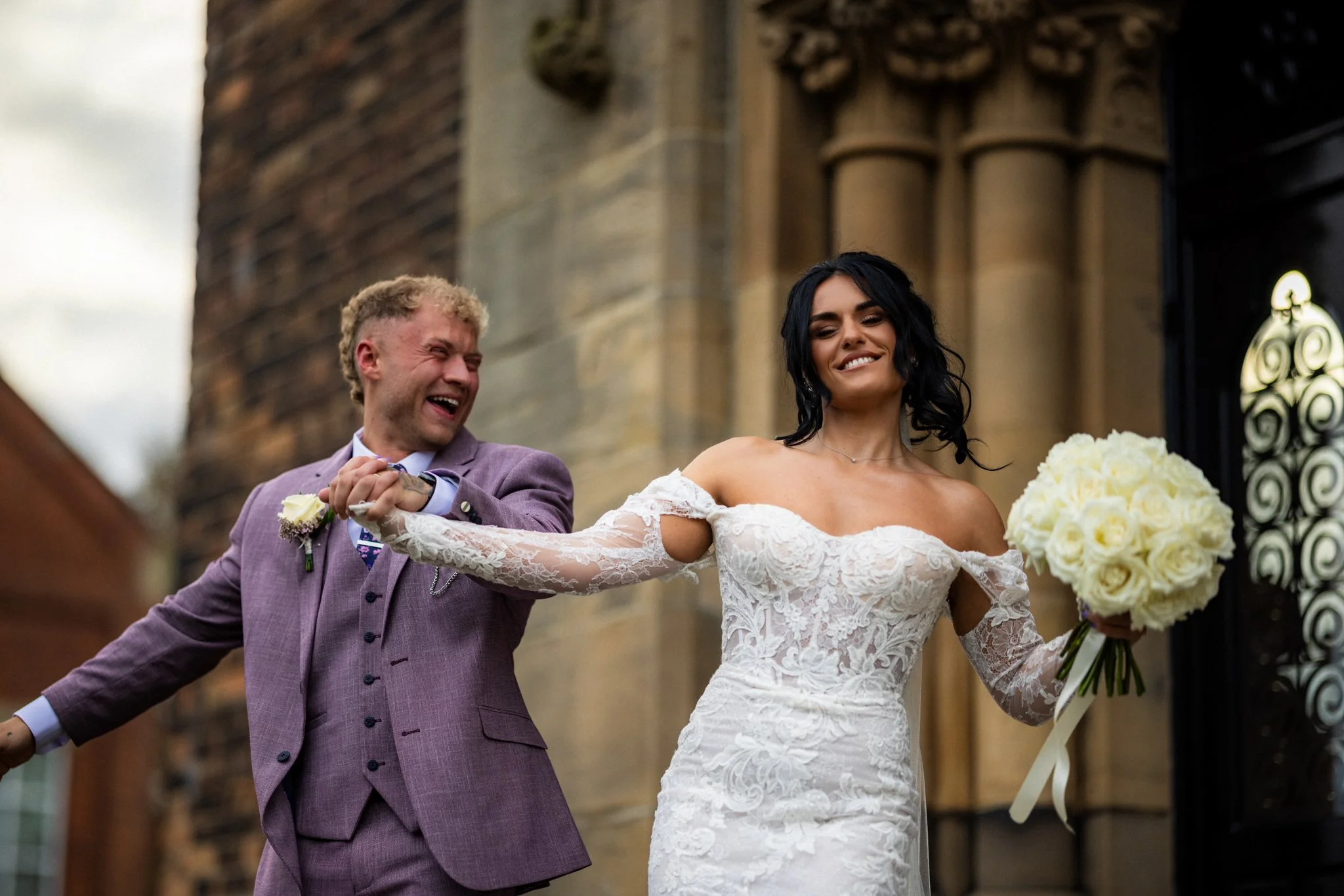 A bride and groom holding hands and smiling outside of a church. The bride is wearing a white lace wedding dress and holding a bouquet of white flowers, and the groom is in a purple suit.