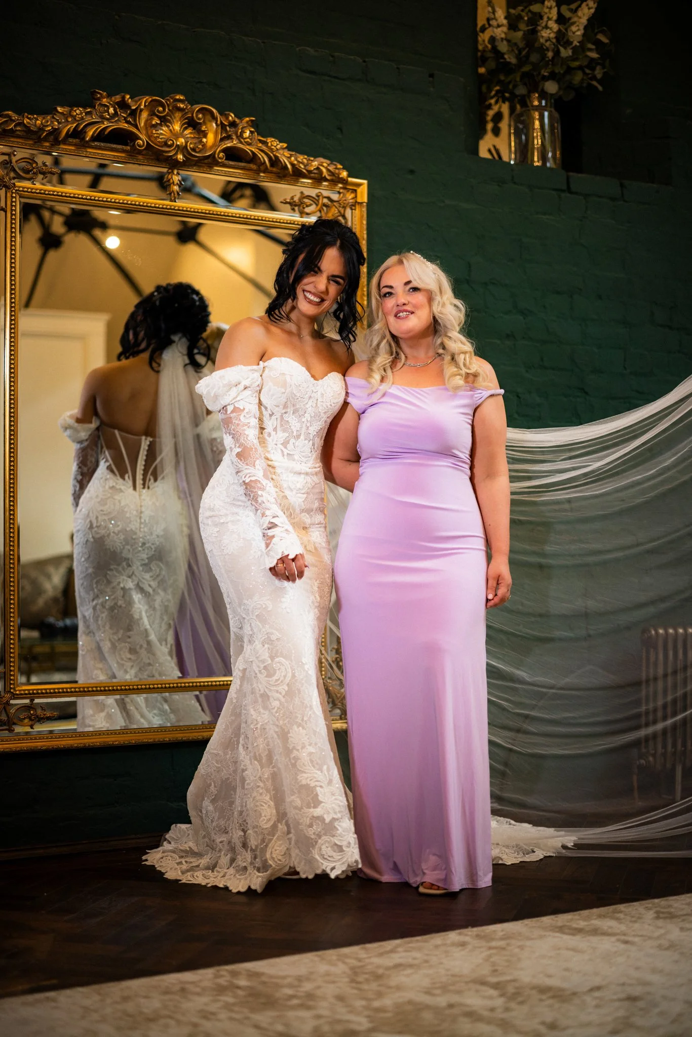 Two women standing in front of a large ornate mirror, with one wearing a white lace bridal gown and the other in a lavender dress, smiling and posing at a wedding or special event.