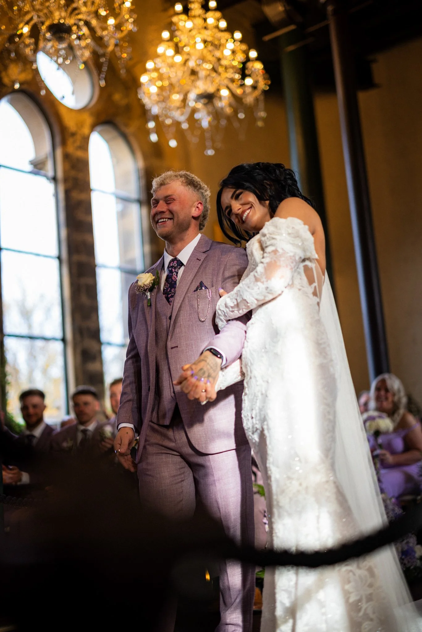 A happy bride and groom holding hands during a wedding ceremony inside a beautifully decorated venue with large arched windows and a grand chandelier ceiling.