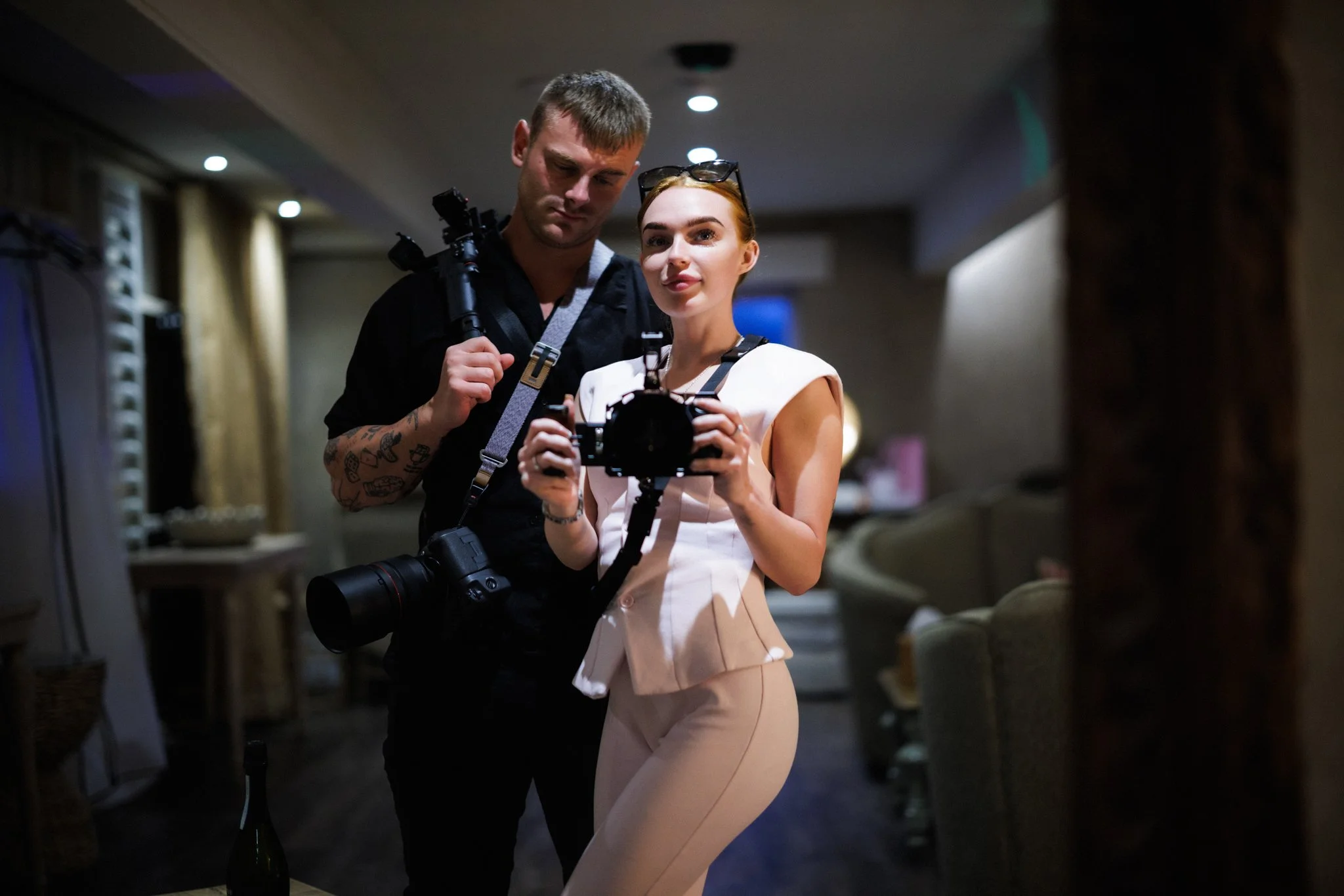 A man and woman in an indoor setting, possibly a conference or event, with the woman holding a camera and the man holding a camera tripod strap, both engaged in an activity together.