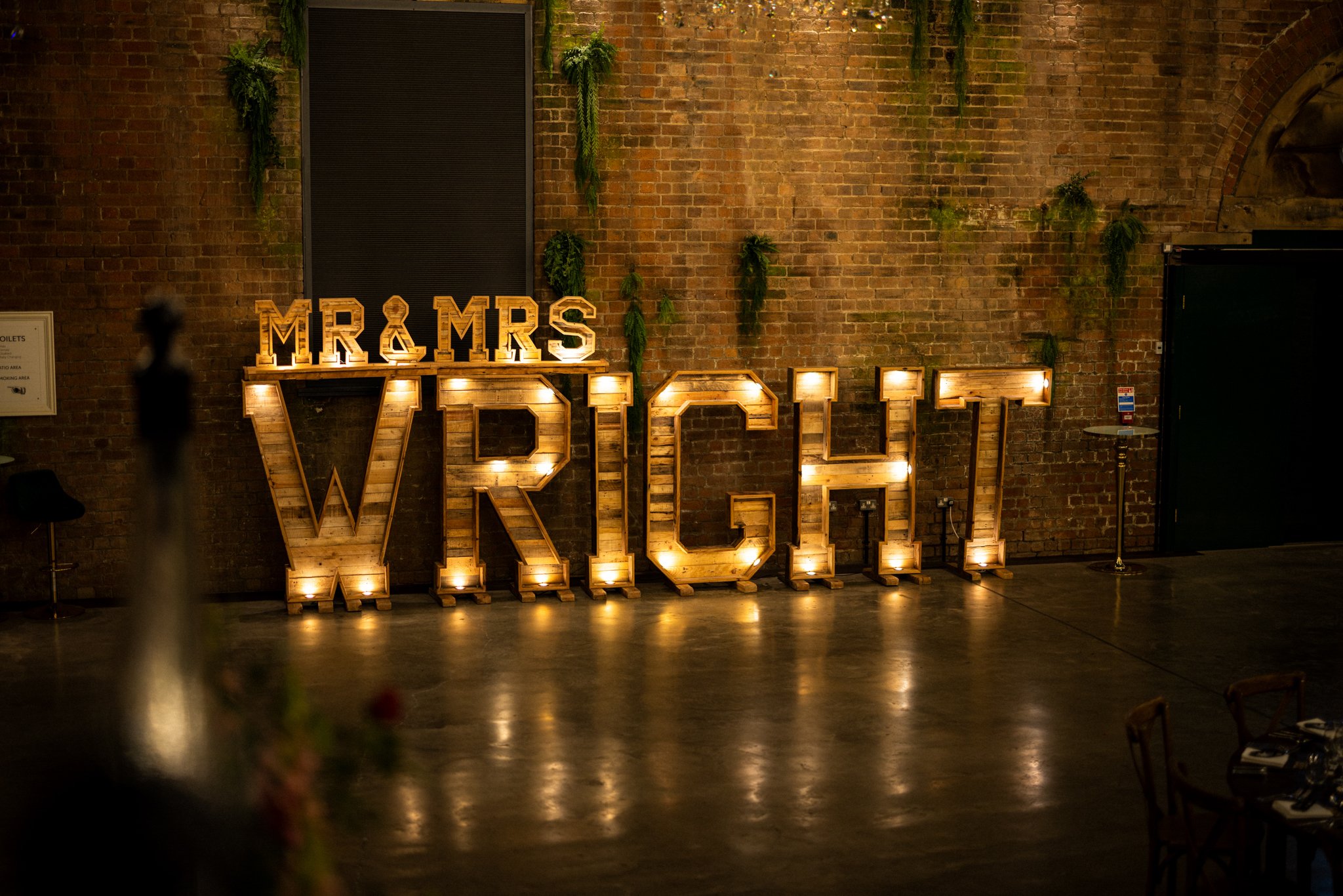 Decorative illuminated wooden letters spelling 'MR & MRS' and 'WEDDING' against a brick wall with hanging plants.