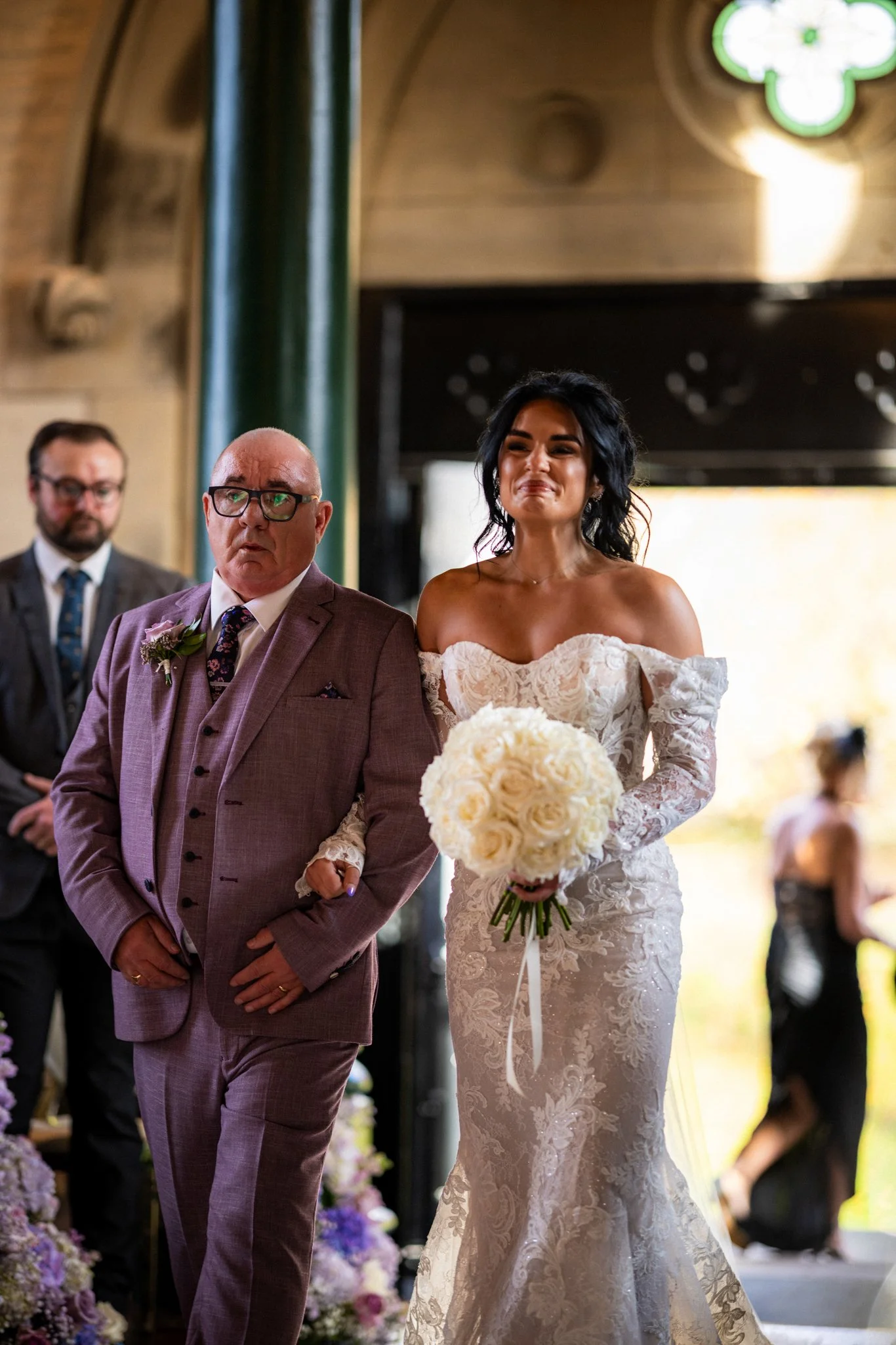 A bride in a lace wedding gown holding a bouquet of white roses walking down the aisle with her father, who is wearing a brown suit, at a wedding ceremony with floral decorations and guests in the background.