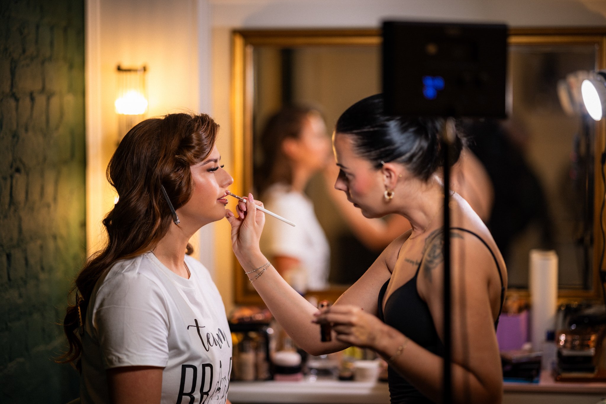 A woman in a white T-shirt with text is having makeup applied to her lips by a makeup artist in a black tank top. They are in a room with warm lighting, a mirror, and other women in the background.