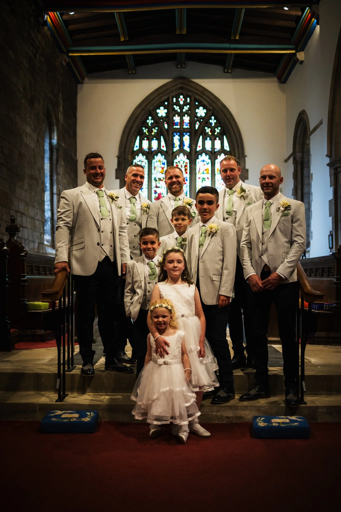 The image depicts a group of people dressed in formal attire, standing inside a church with stained glass windows behind them. The group appears to be celebrating a wedding or special occasion.