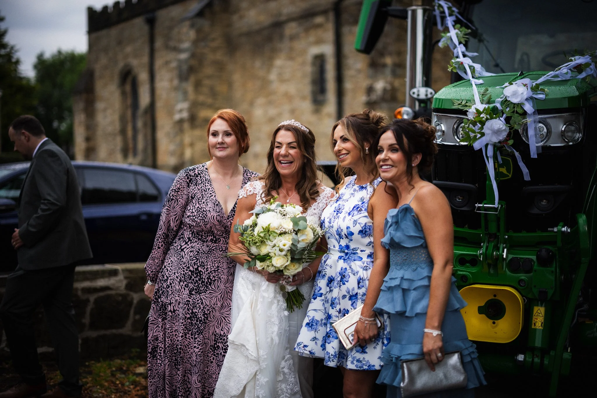Group of four women dressed in formal attire, standing outdoors near a vintage stone building and a decorated green vehicle, celebrating a wedding with a bouquet and smiling faces.